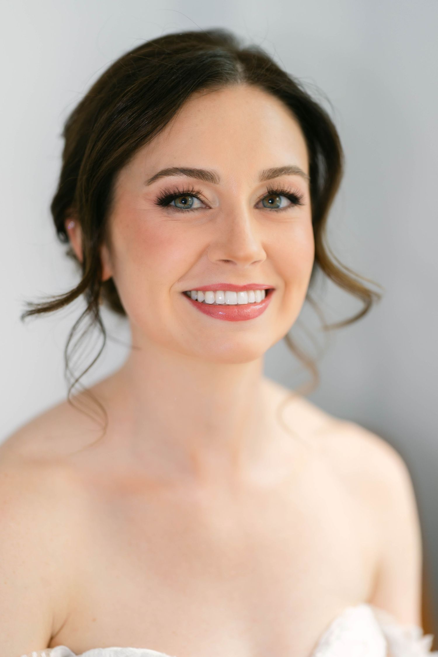 Smiling bride with elegant updo, rosy makeup, and strapless white gown posing for a bridal portrait.
