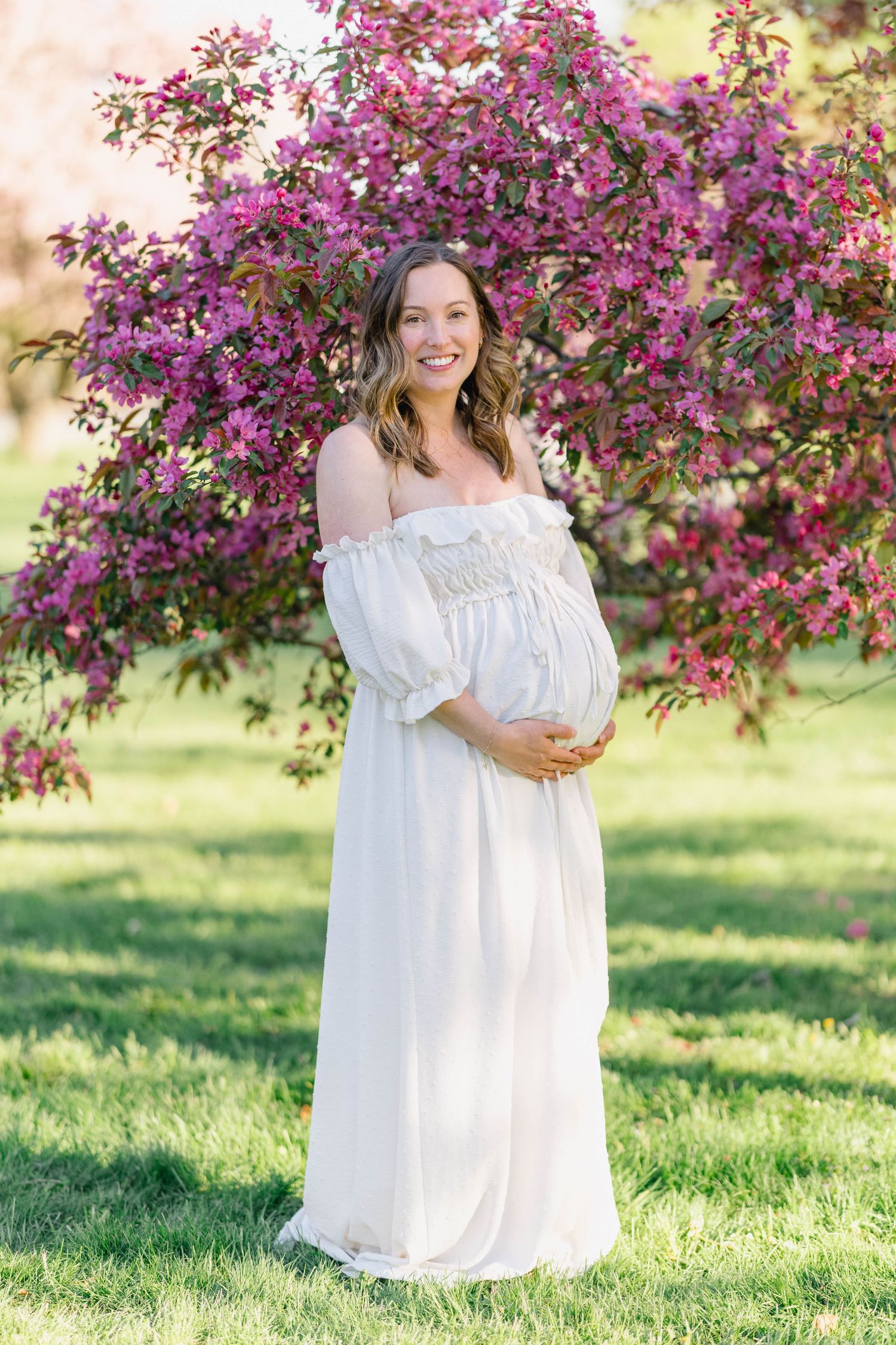 Smiling pregnant woman in white off-shoulder dress cradling baby bump near blooming pink flowering tree outdoors.