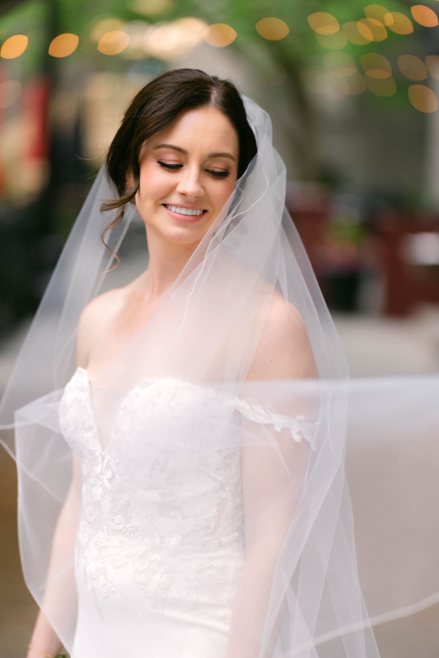 Smiling bride in off-shoulder lace gown with flowing white veil draped around her outdoors.