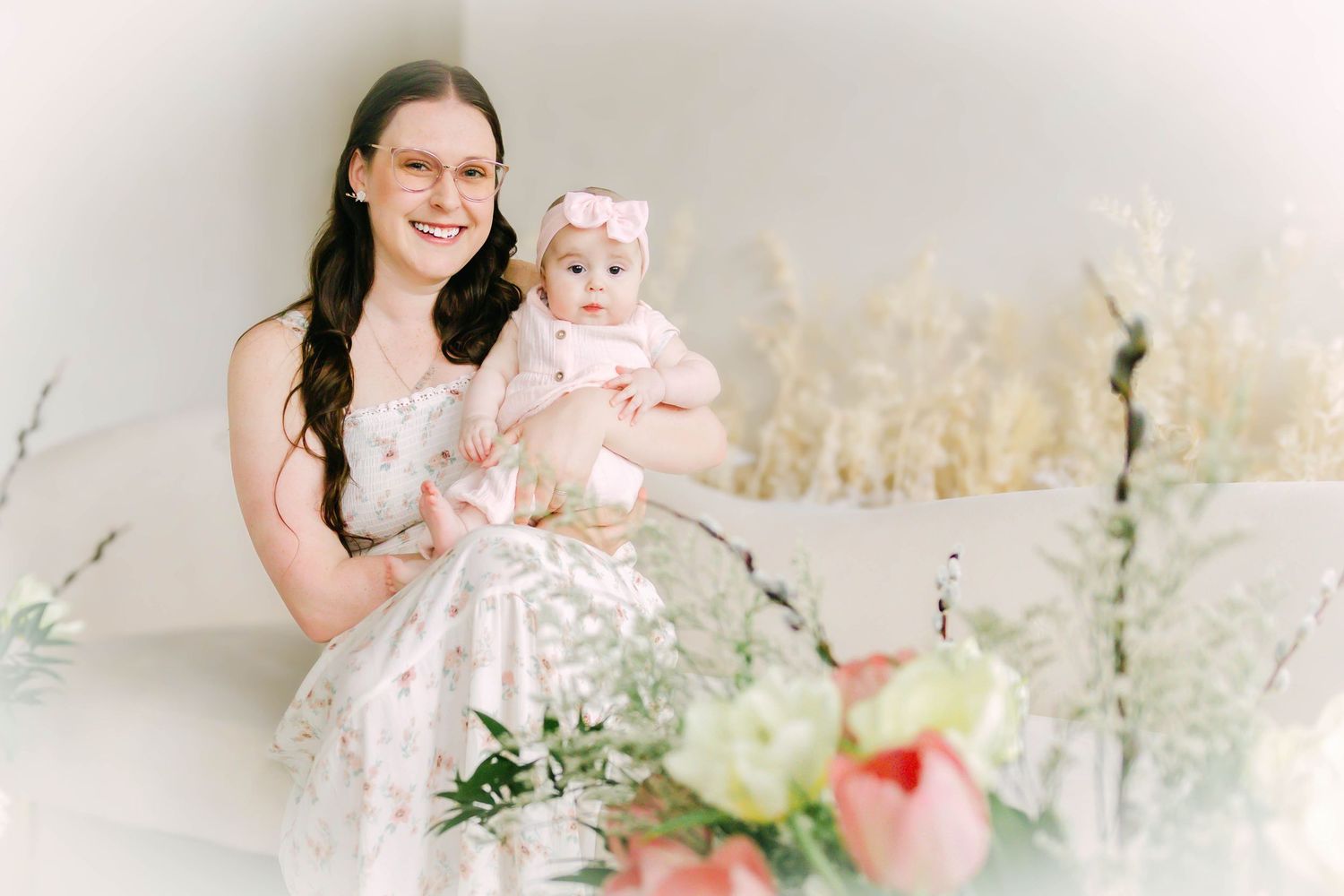 A woman in a floral dress holds a baby wearing a pink outfit against a light background with soft floral decorations.