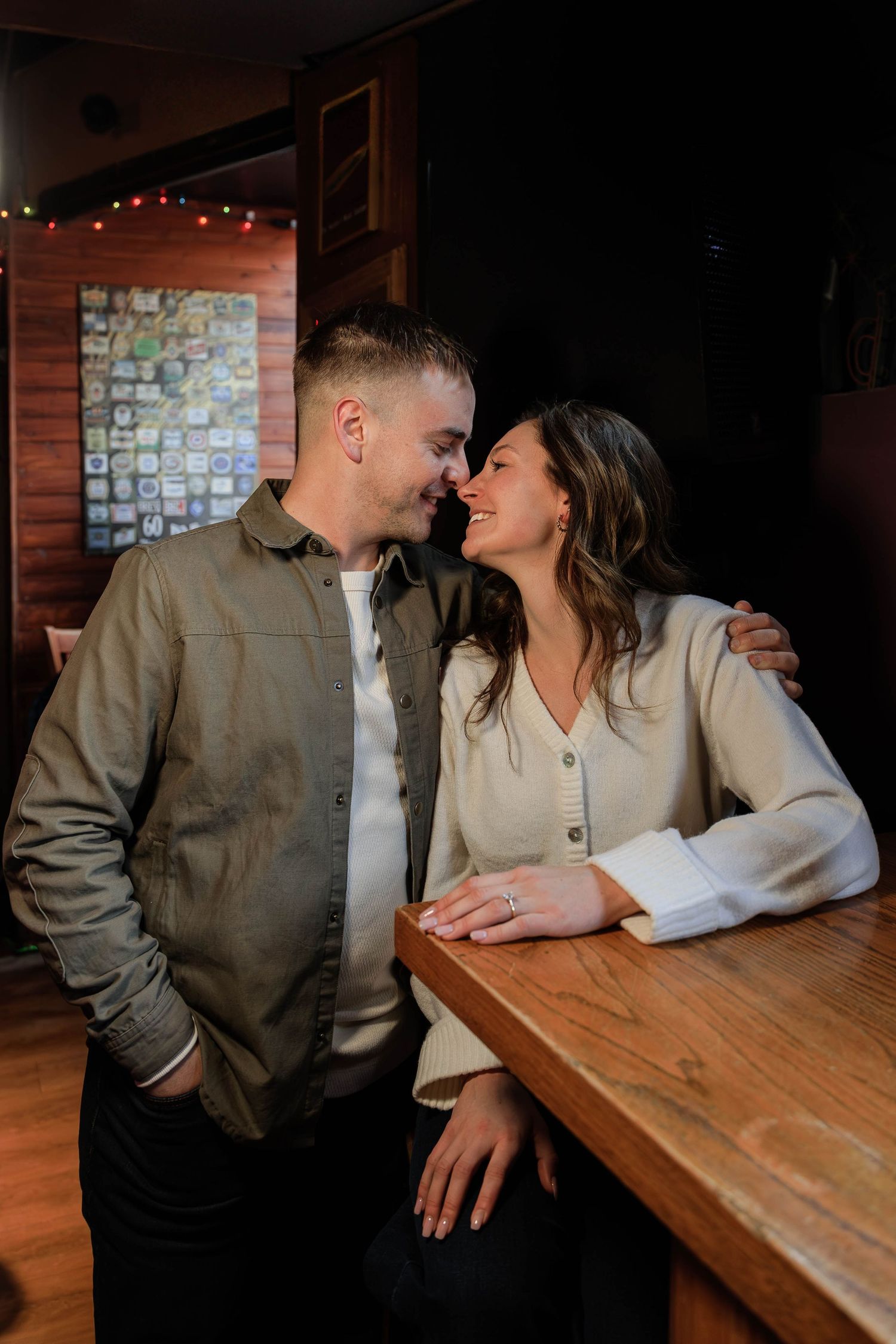 A romantic couple shares an intimate moment at a wooden bar table under warm lighting with string lights in the background.