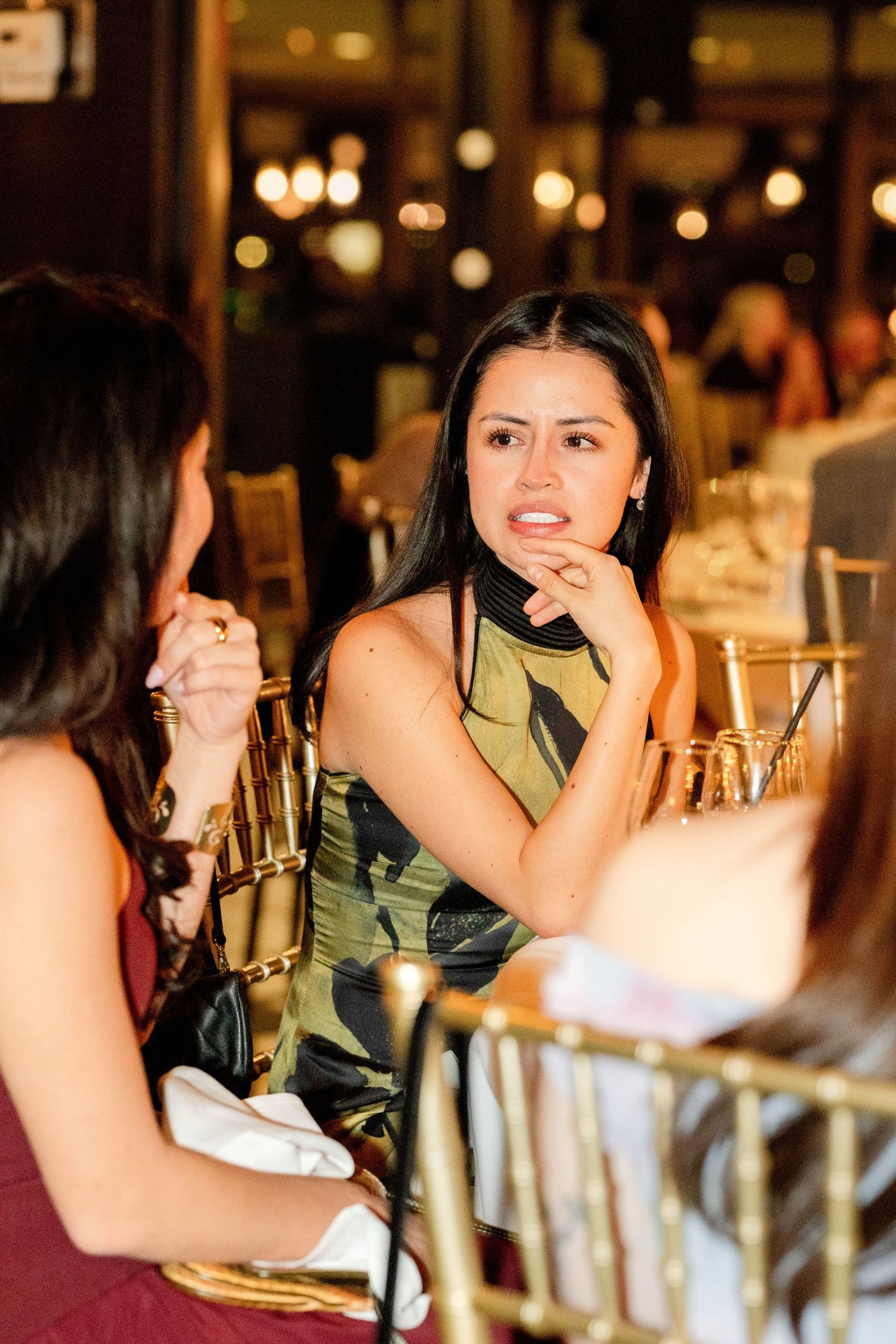 Woman in green floral dress sits at elegant gold-chaired restaurant table, resting chin on hand during conversation.