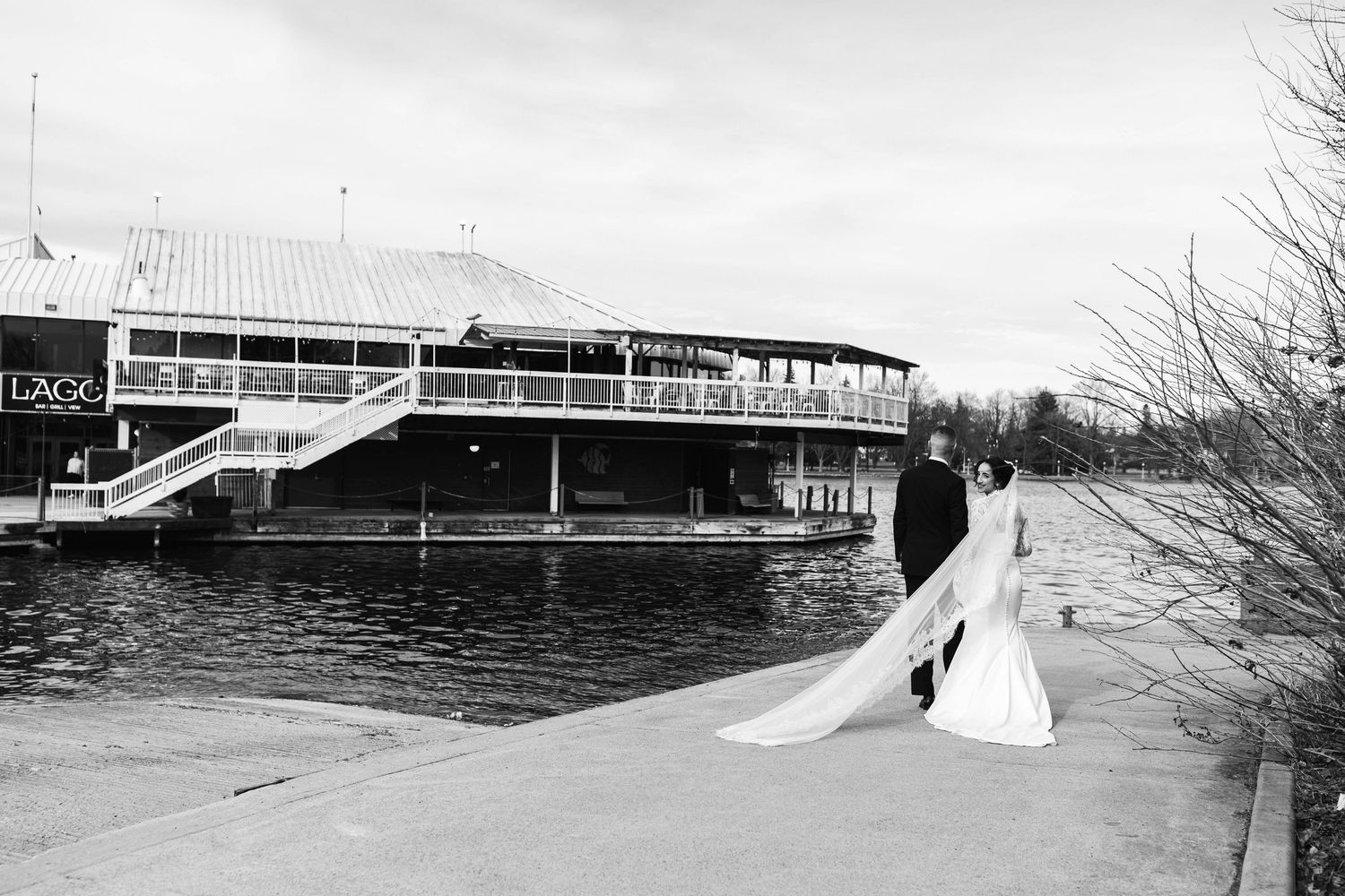 Black and white photo of a bride and groom posing by a lakeside venue with a large waterfront building in the background.