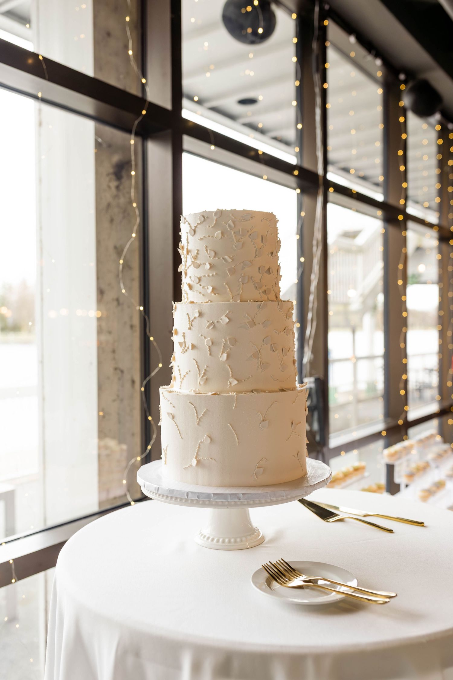 Three-tier ivory wedding cake with textured butterfly decorations on a white stand, gold cutlery beside large windows.