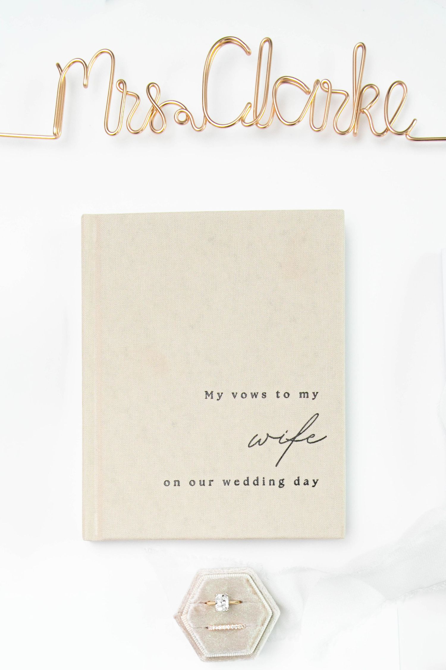 Wedding flat lay featuring a vow book, diamond ring, and gold wire Mrs. Clarke name sign on white background.