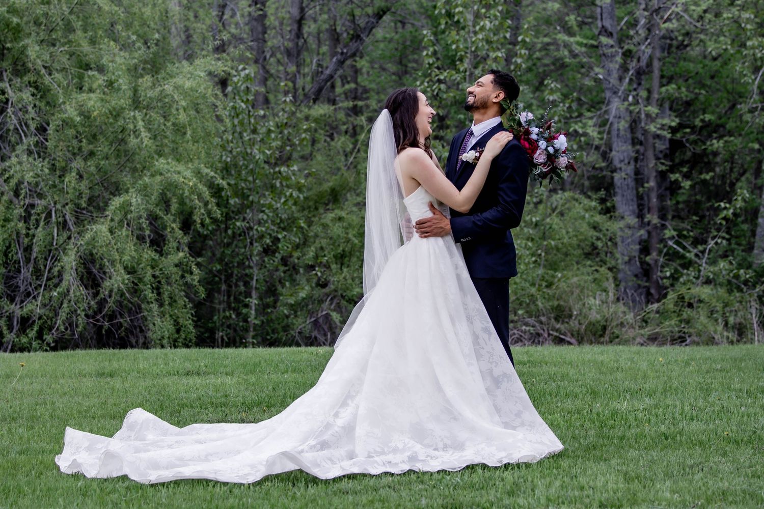 A couple in wedding attire embraces on a lush green lawn with forest trees in the background.