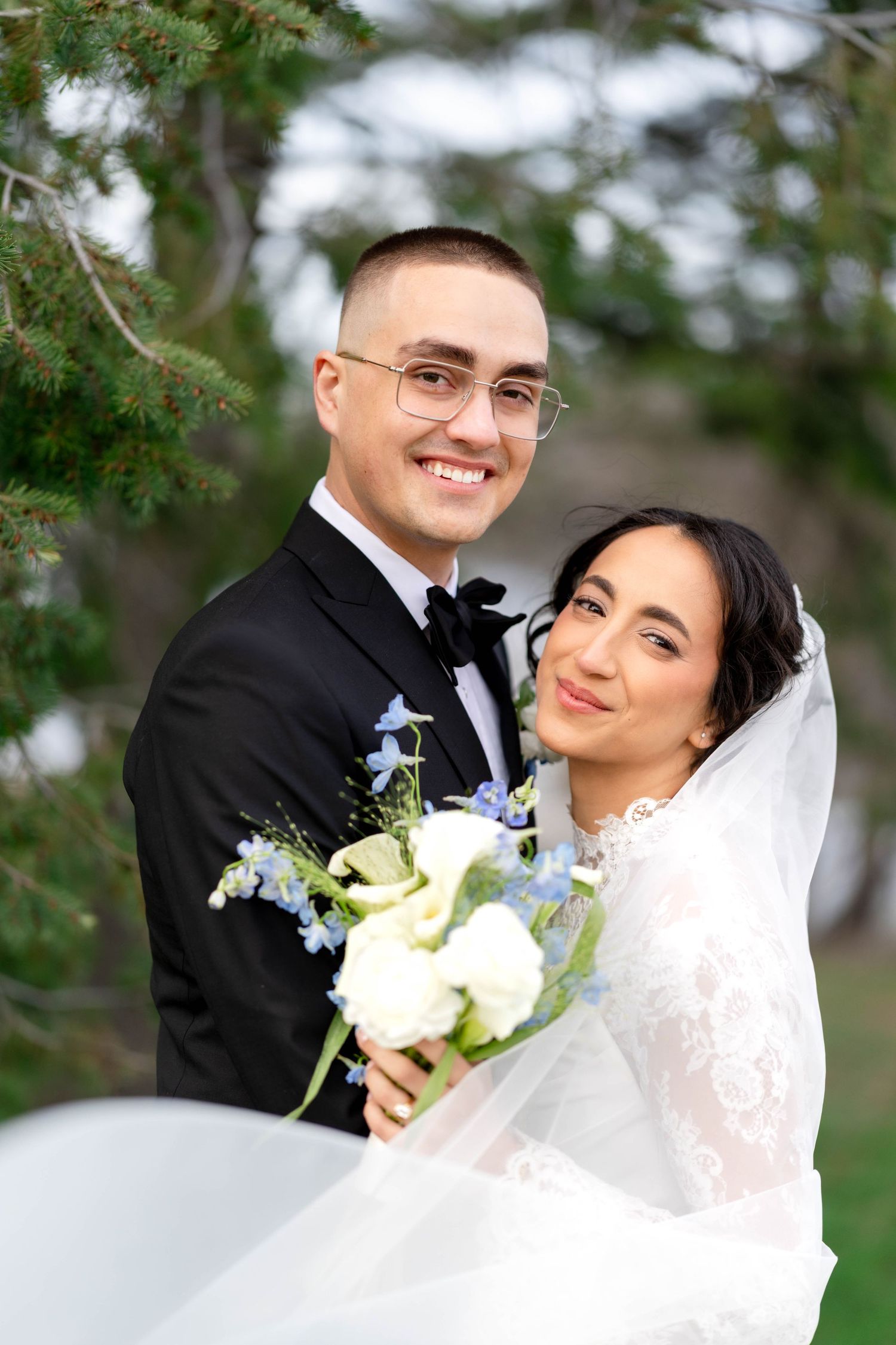 Smiling bride and groom pose outdoors holding white and blue bouquet, groom in black tuxedo, bride in lace wedding dress with veil.