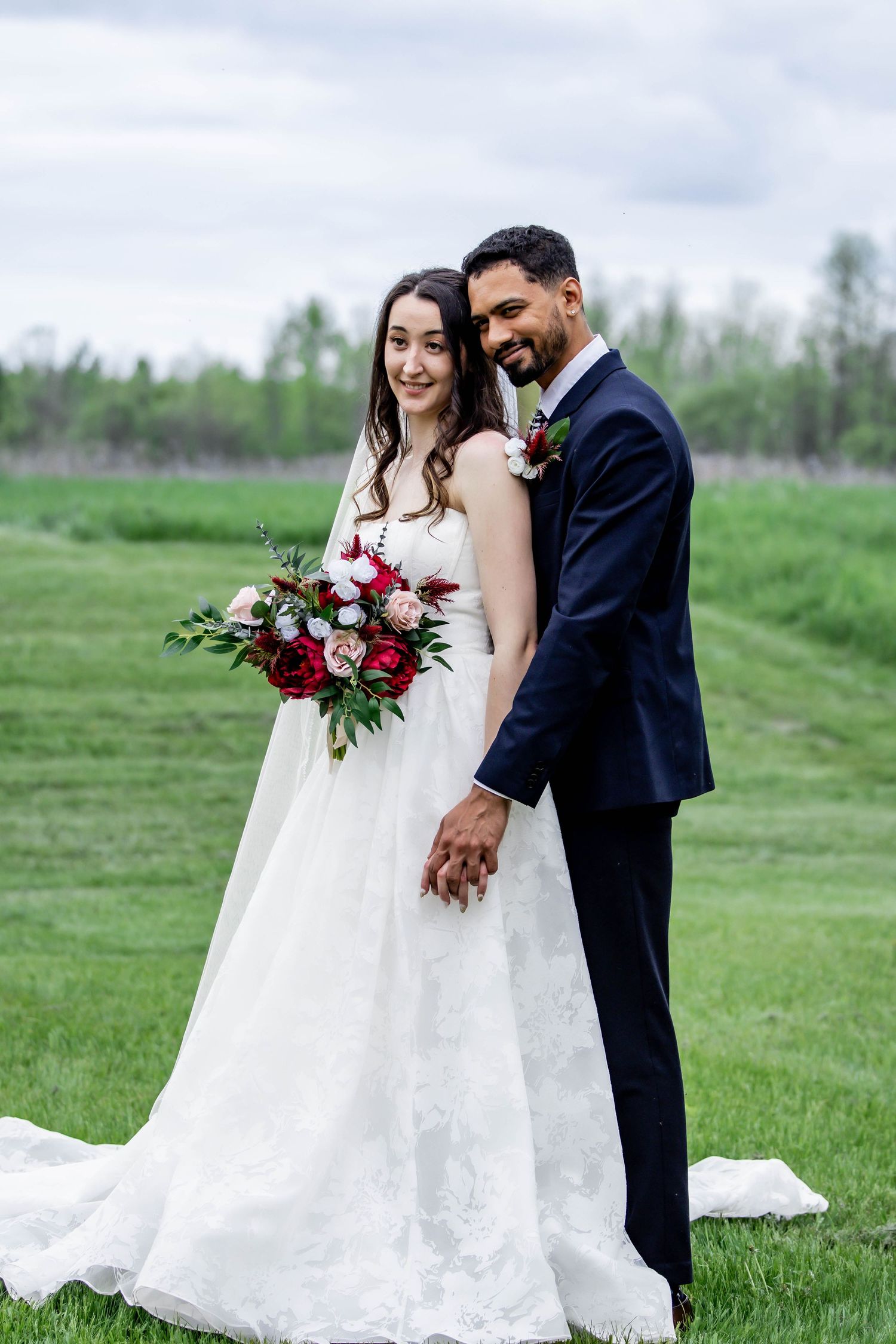 A couple shares an intimate moment in a grassy field during their wedding photoshoot, with the bride holding a red bouquet.