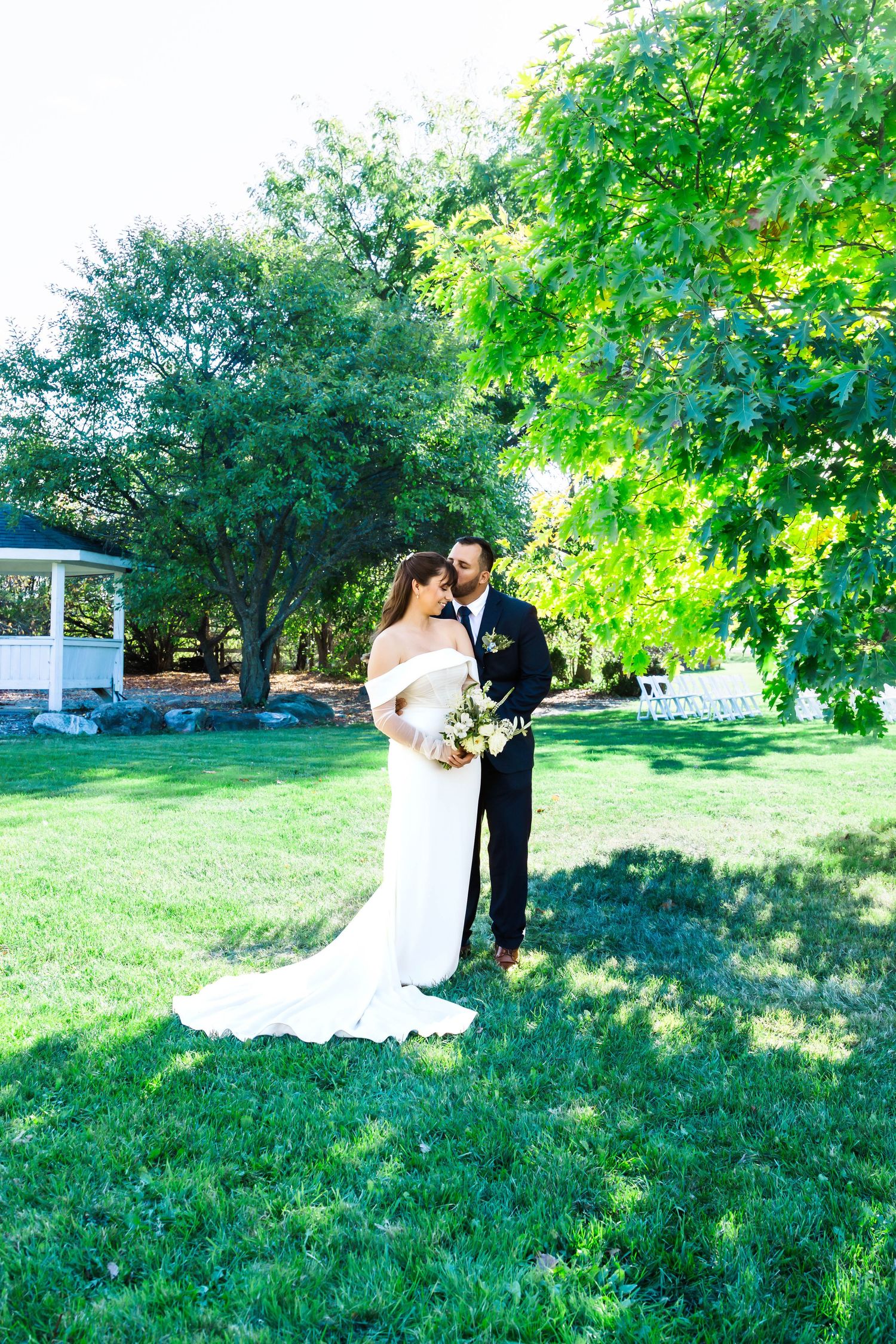 A couple poses on a sunny lawn with a white gazebo and lush trees in the background on their wedding day.