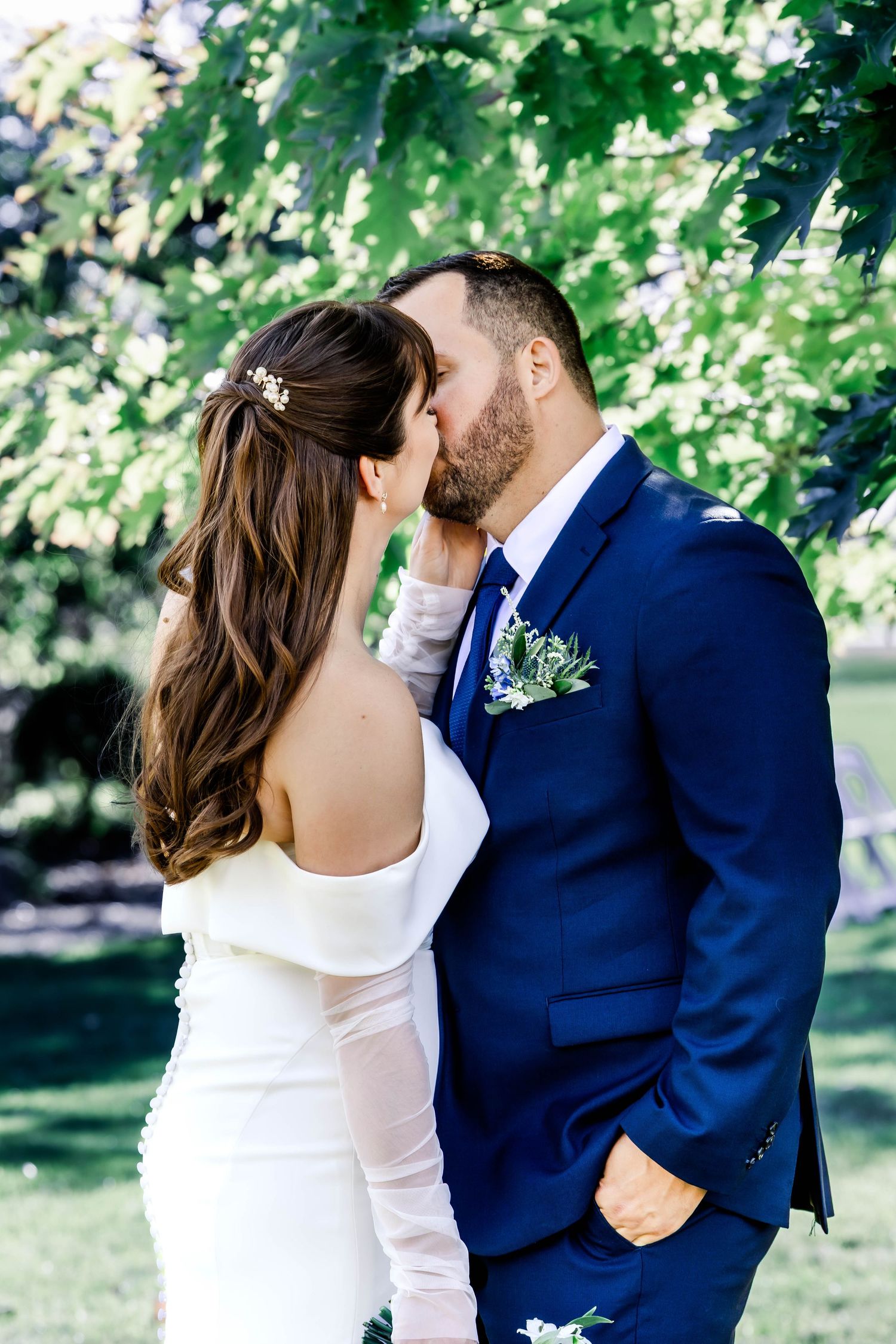 A couple in wedding attire share an intimate moment outdoors under green trees on their special day.