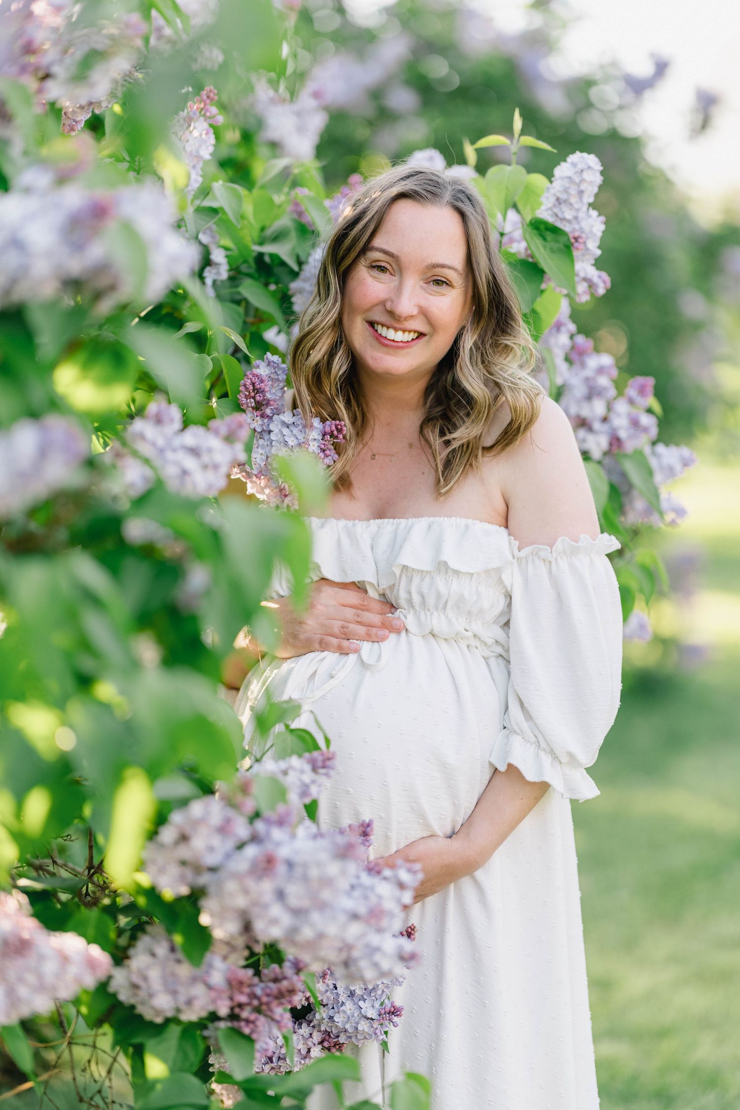 Smiling pregnant woman in white off-shoulder dress cradling baby bump among blooming lilac bushes outdoors.