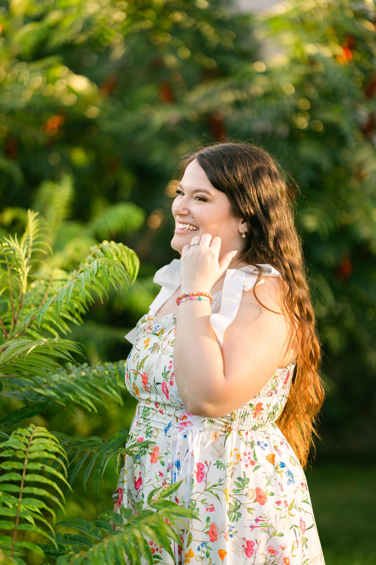 Smiling young woman in floral sundress standing among lush green ferns and plants outdoors in golden sunlight.