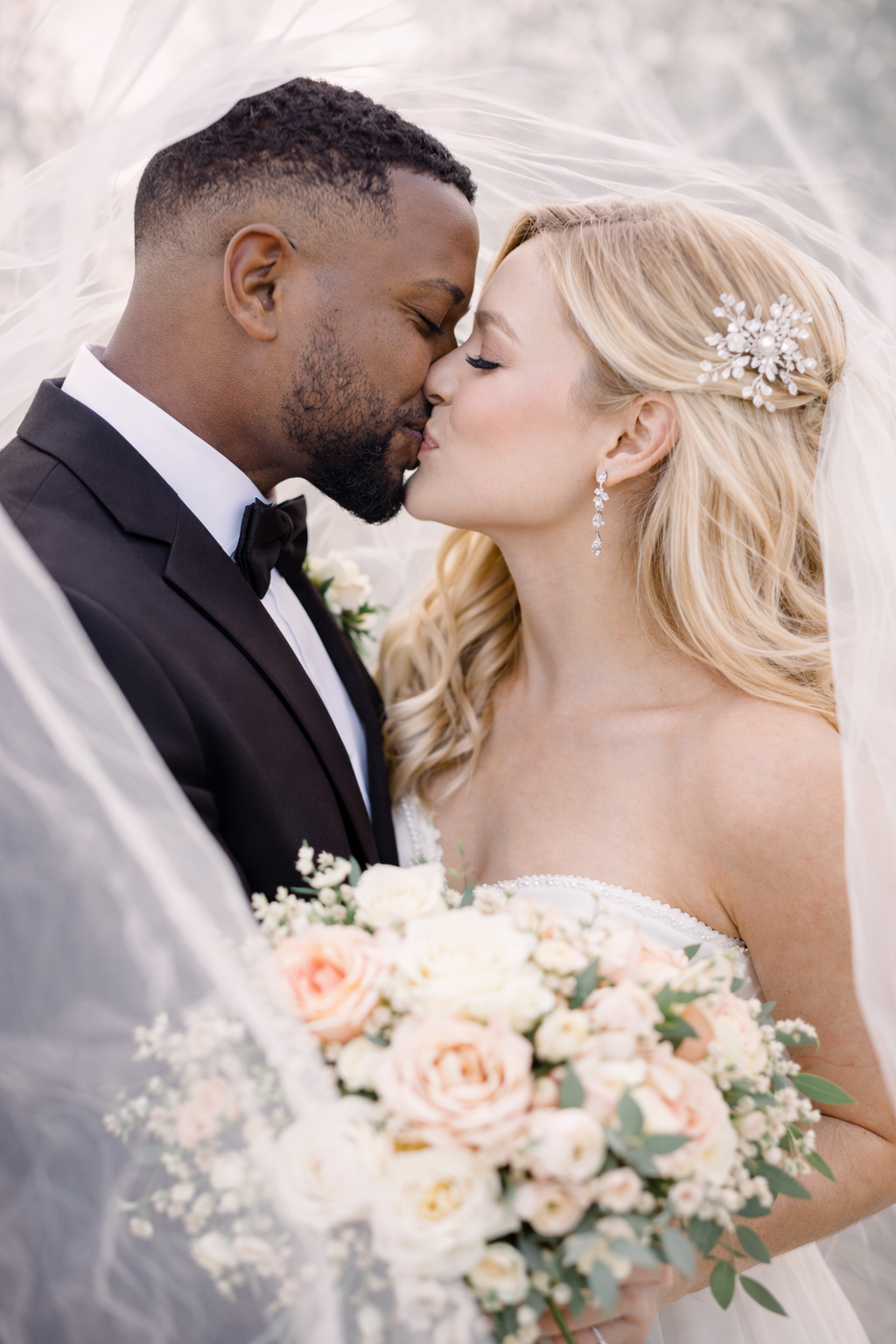 A romantic wedding portrait with a couple sharing a tender kiss under a veil while holding a peach and white rose bouquet.