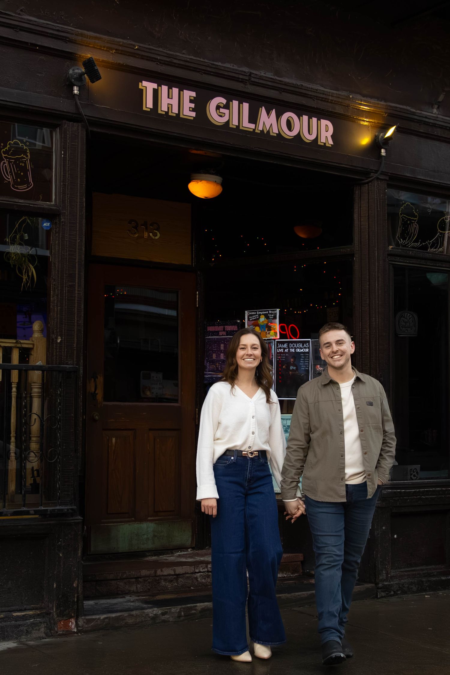 A couple poses together outside The Gilmour establishment at night with illuminated signage visible.