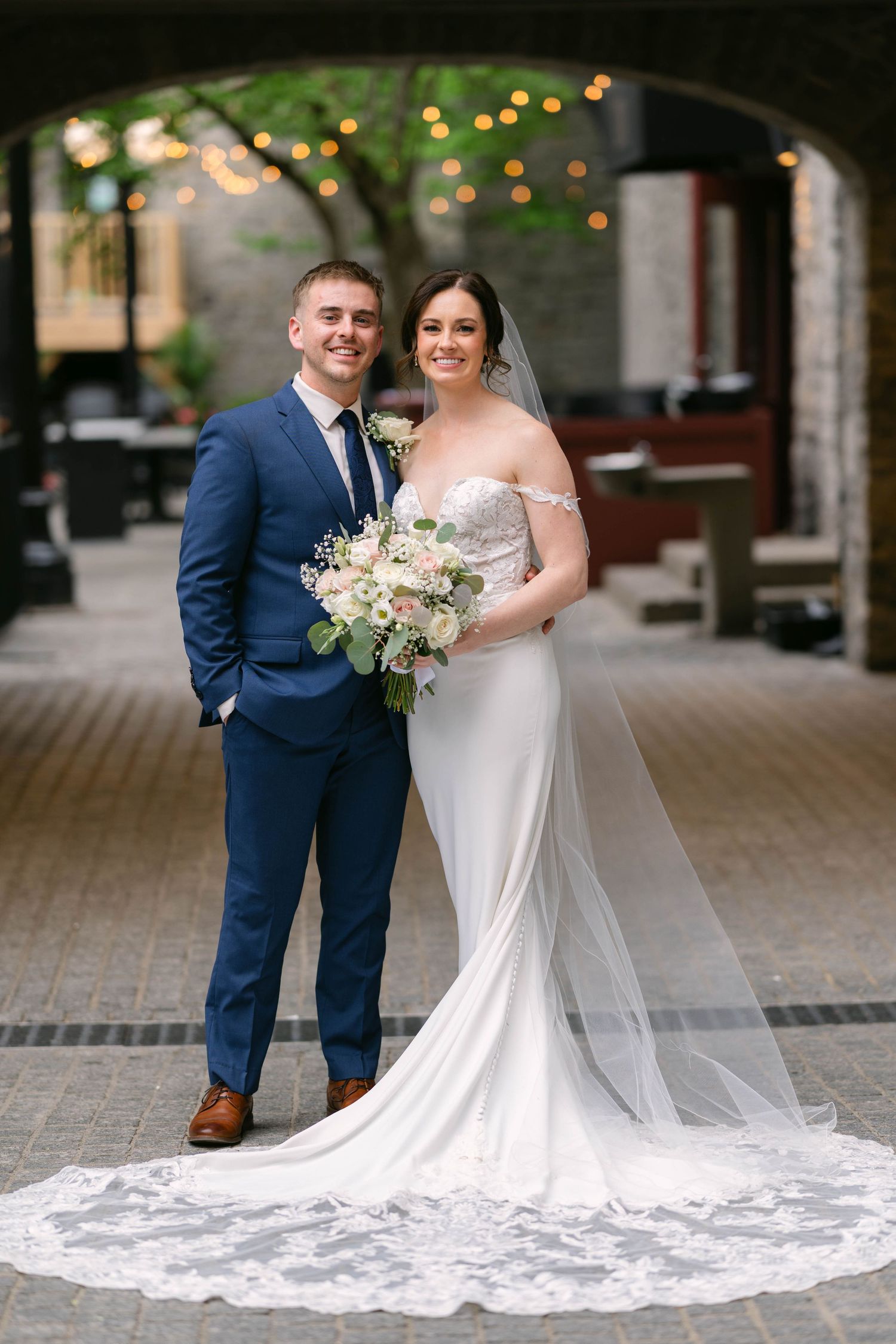 Elegant bride and groom portrait under stone archway, bride holding blush and white floral bouquet.