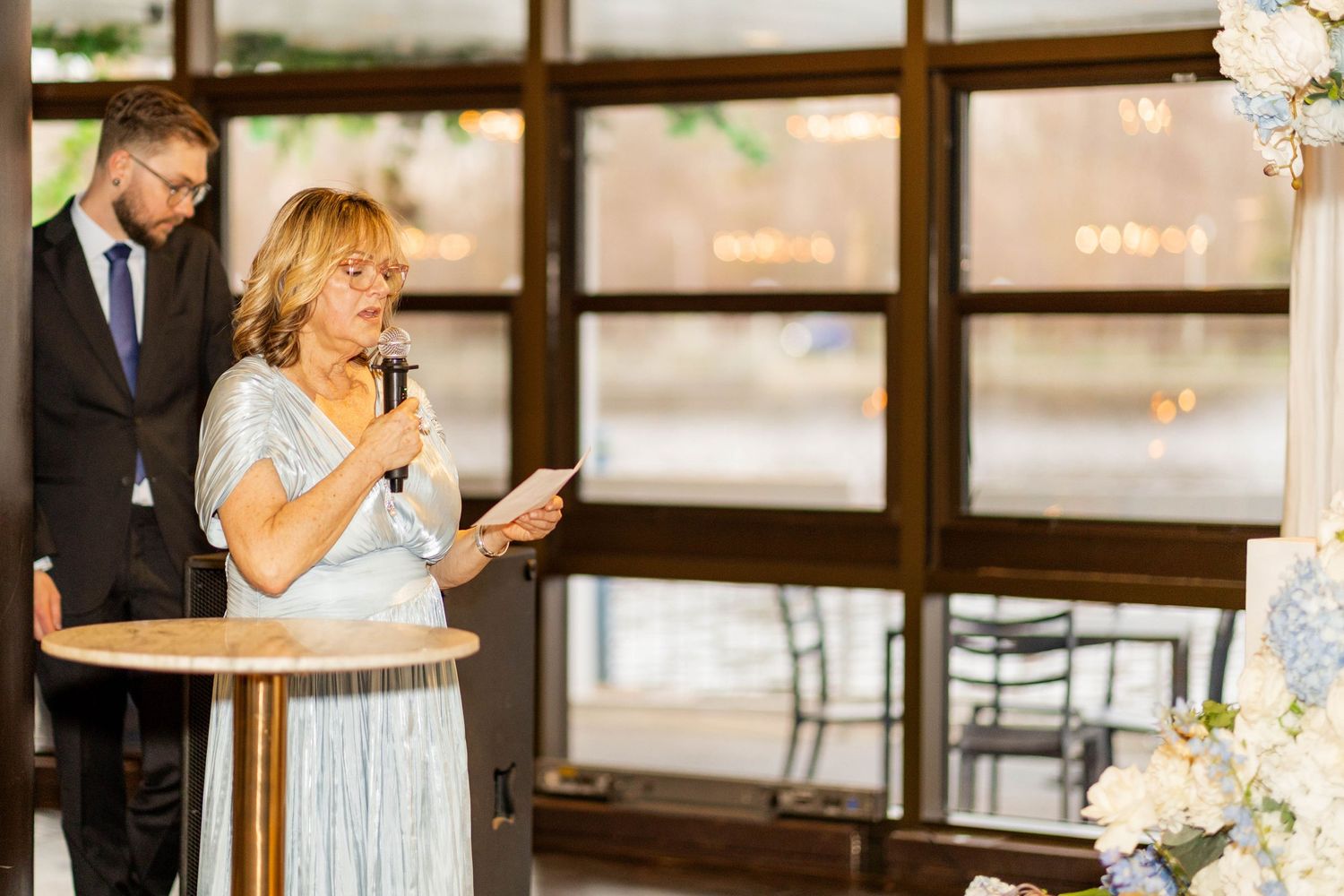Woman in silver dress giving a speech at an elegant wedding reception with floral arrangements.