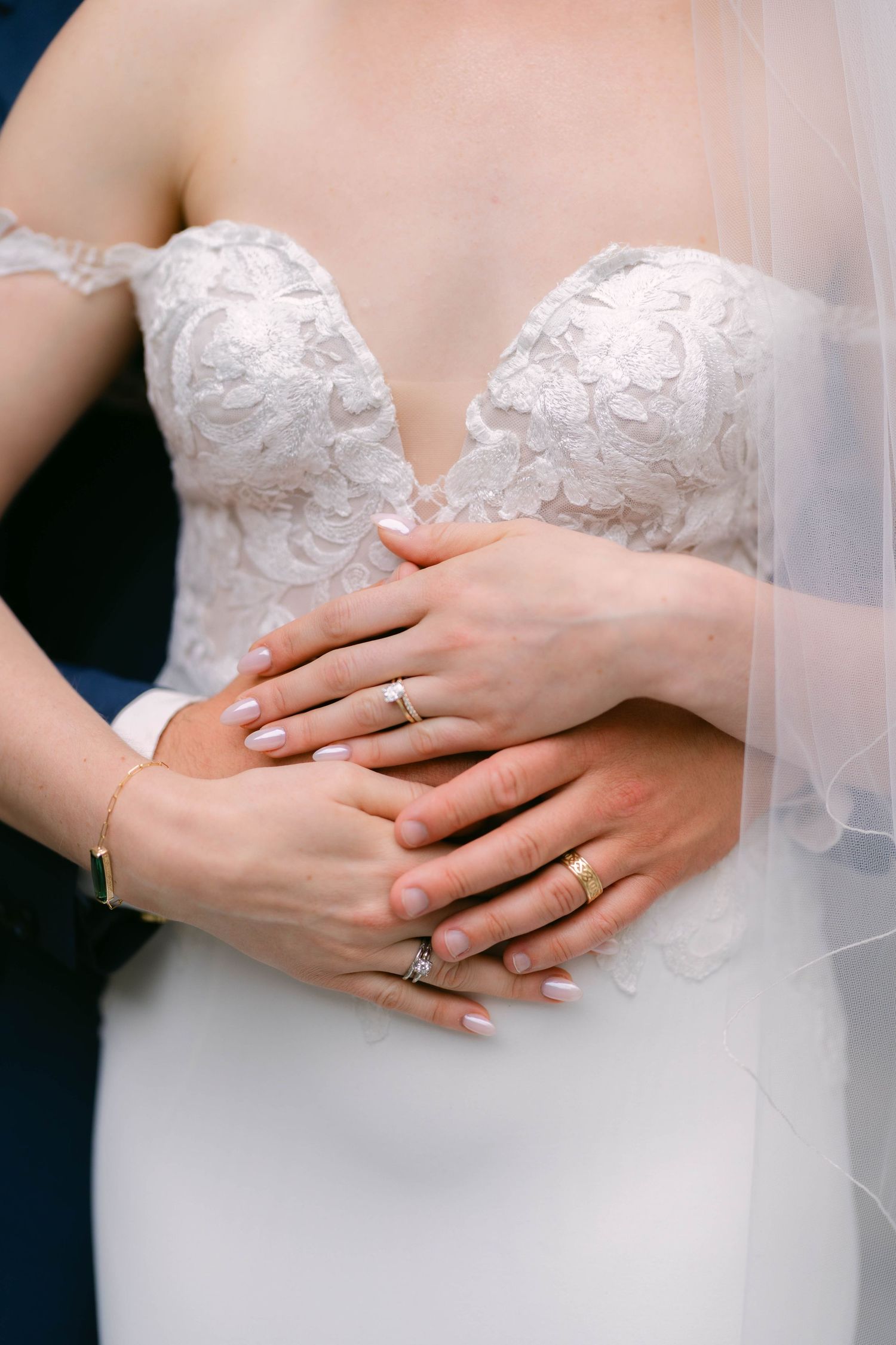 Bride and groom hands with wedding rings clasped together over white lace bridal gown.