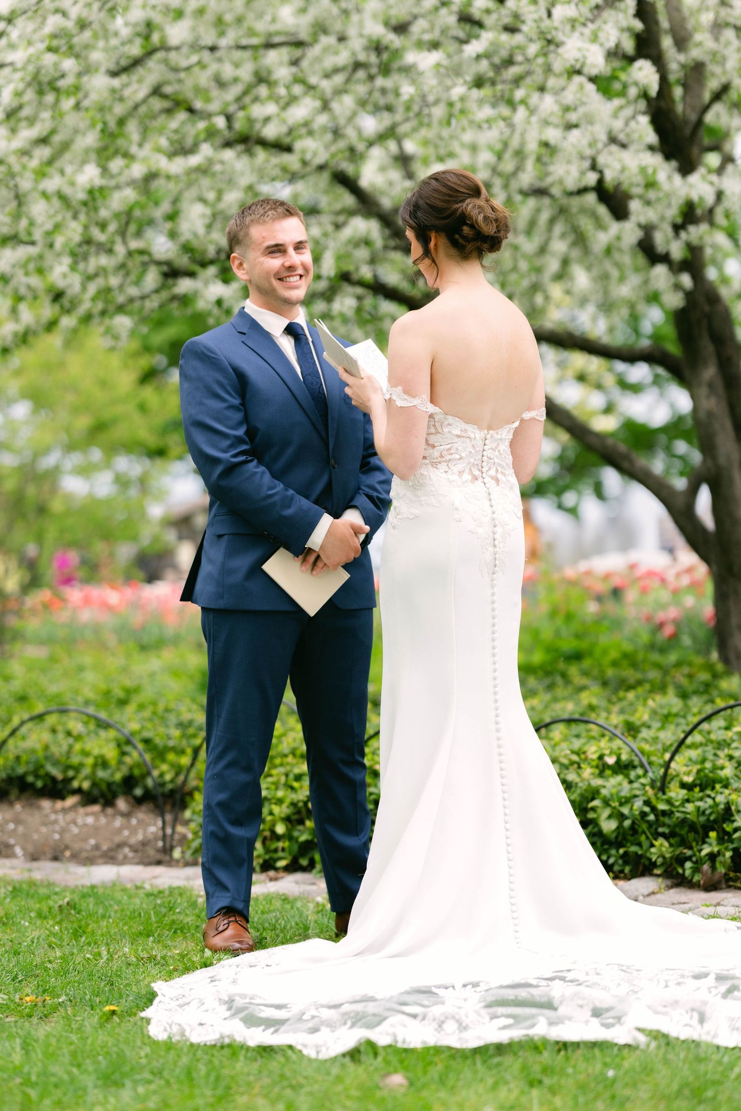 Smiling groom in navy suit exchanges vows with bride in lace wedding dress under blooming tree.