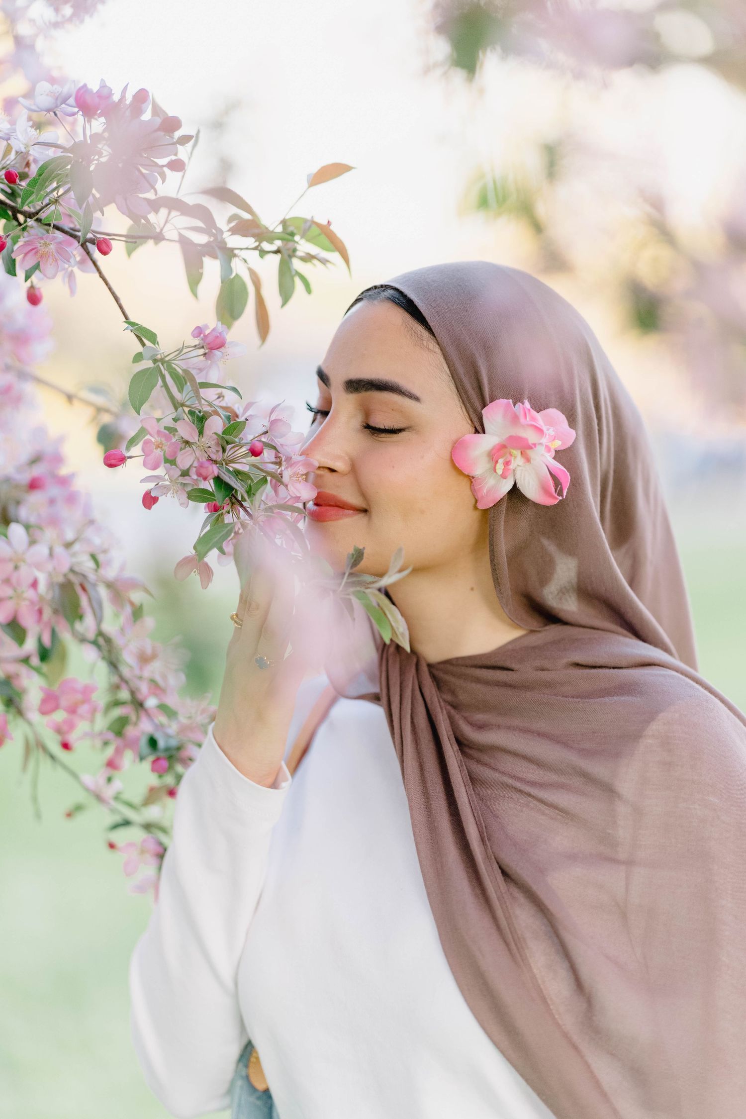 Young Muslim woman in hijab smelling pink cherry blossoms outdoors with eyes closed in serene spring setting.