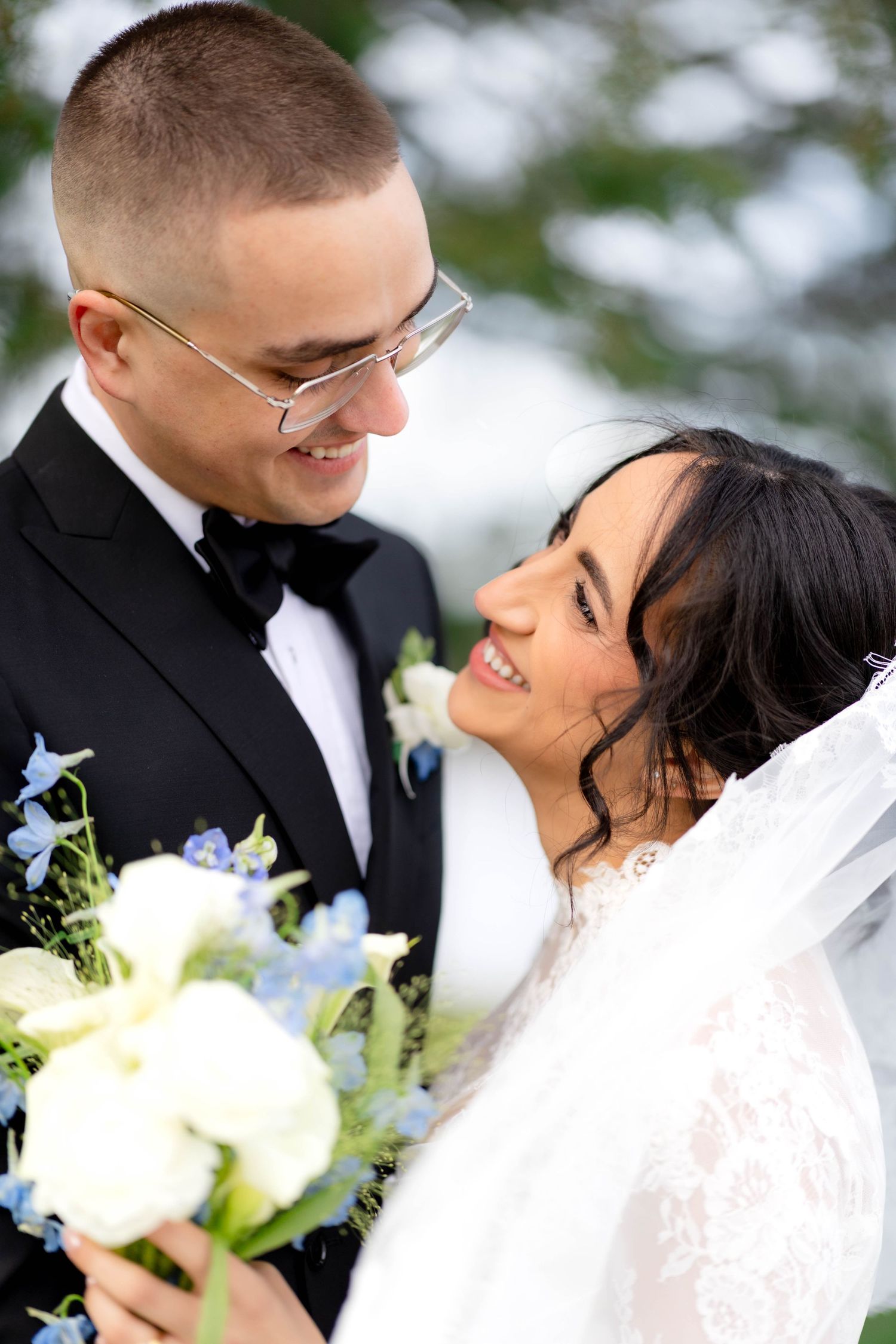 Smiling bride and groom in tuxedo share a romantic moment, bride holding white and blue wedding bouquet.