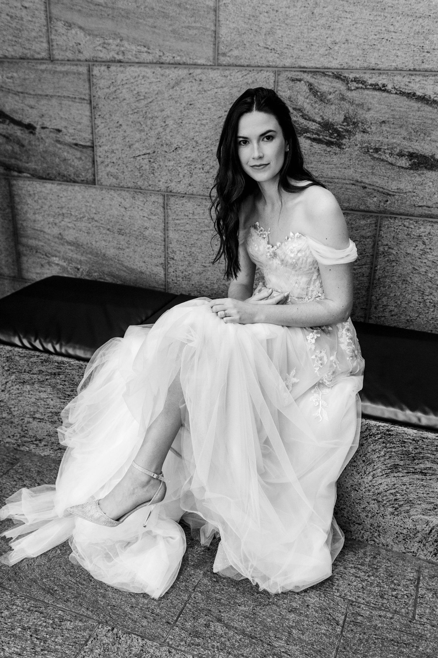 Black and white portrait of someone in an elegant off-shoulder wedding gown sitting on stone steps.