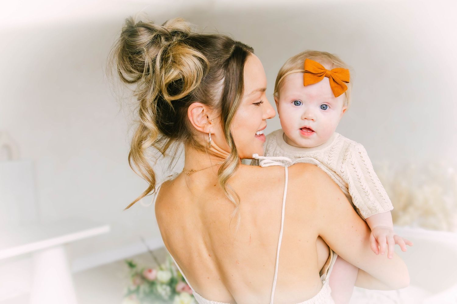 A tender moment captured between a woman in a backless dress and a baby wearing an orange bow headband.