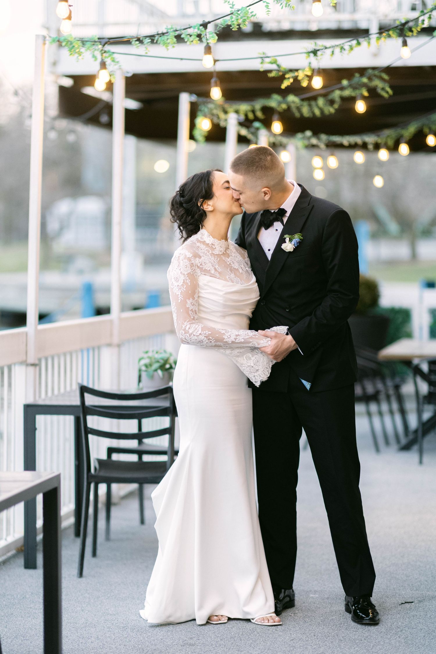 Bride and groom share a romantic kiss on an outdoor patio decorated with string lights and greenery.