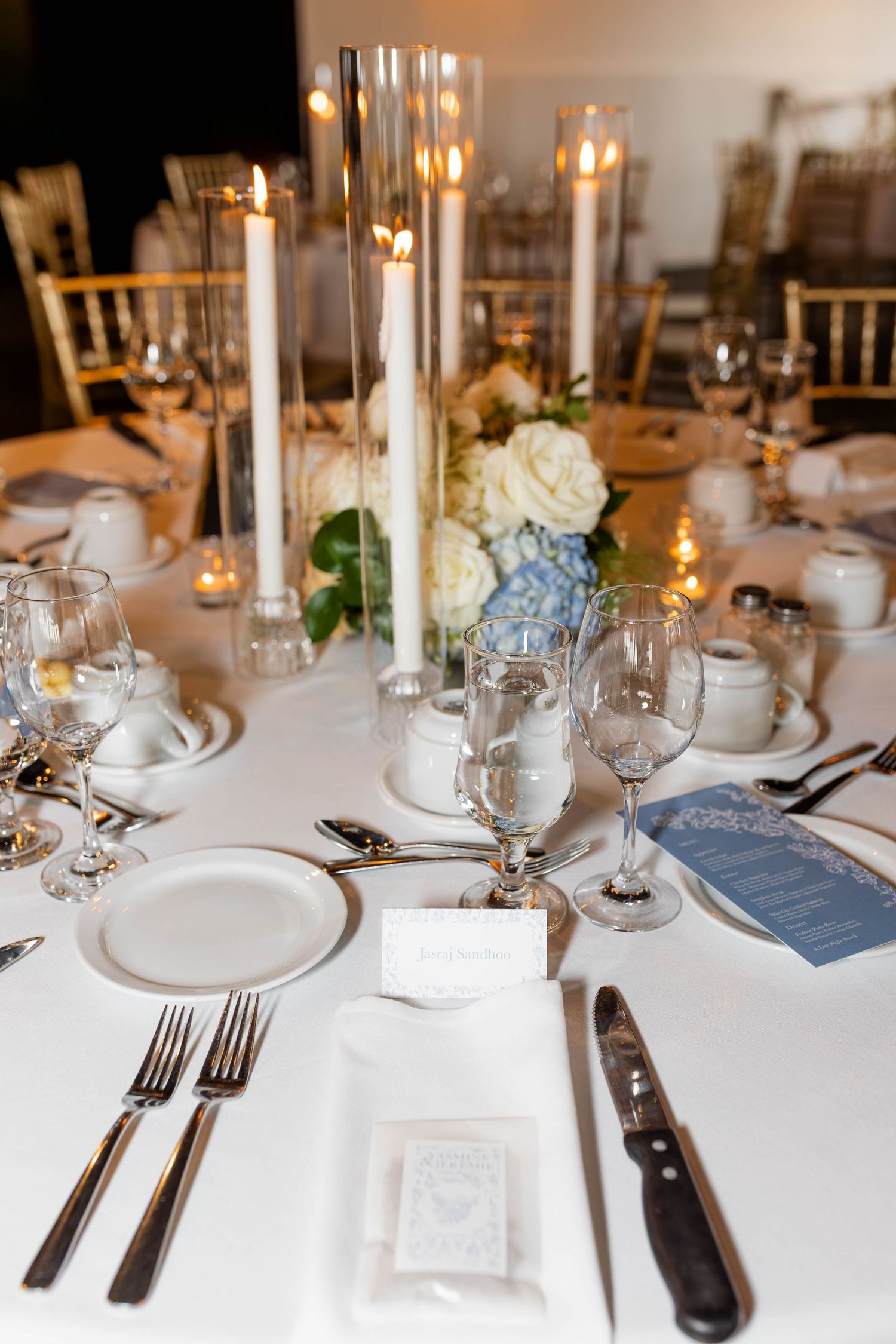 Elegant wedding reception table with white candles, blue and white floral centerpiece, crystal glasses, and formal place settings.