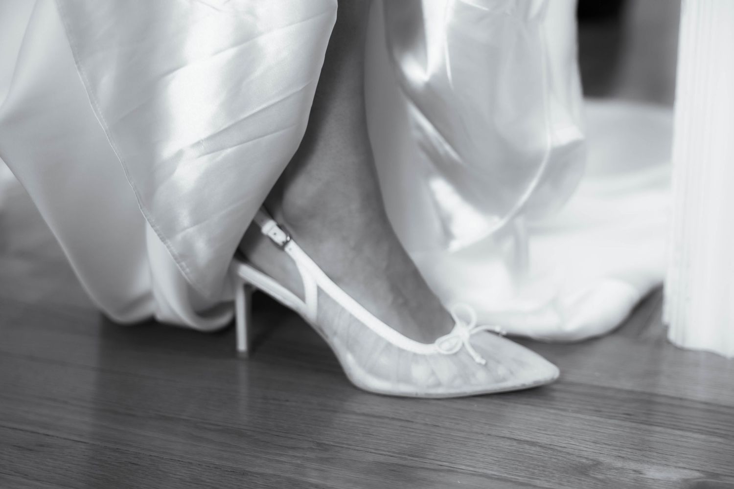 Black and white photo of a bride wearing elegant lace slingback heels beneath a flowing white wedding gown.