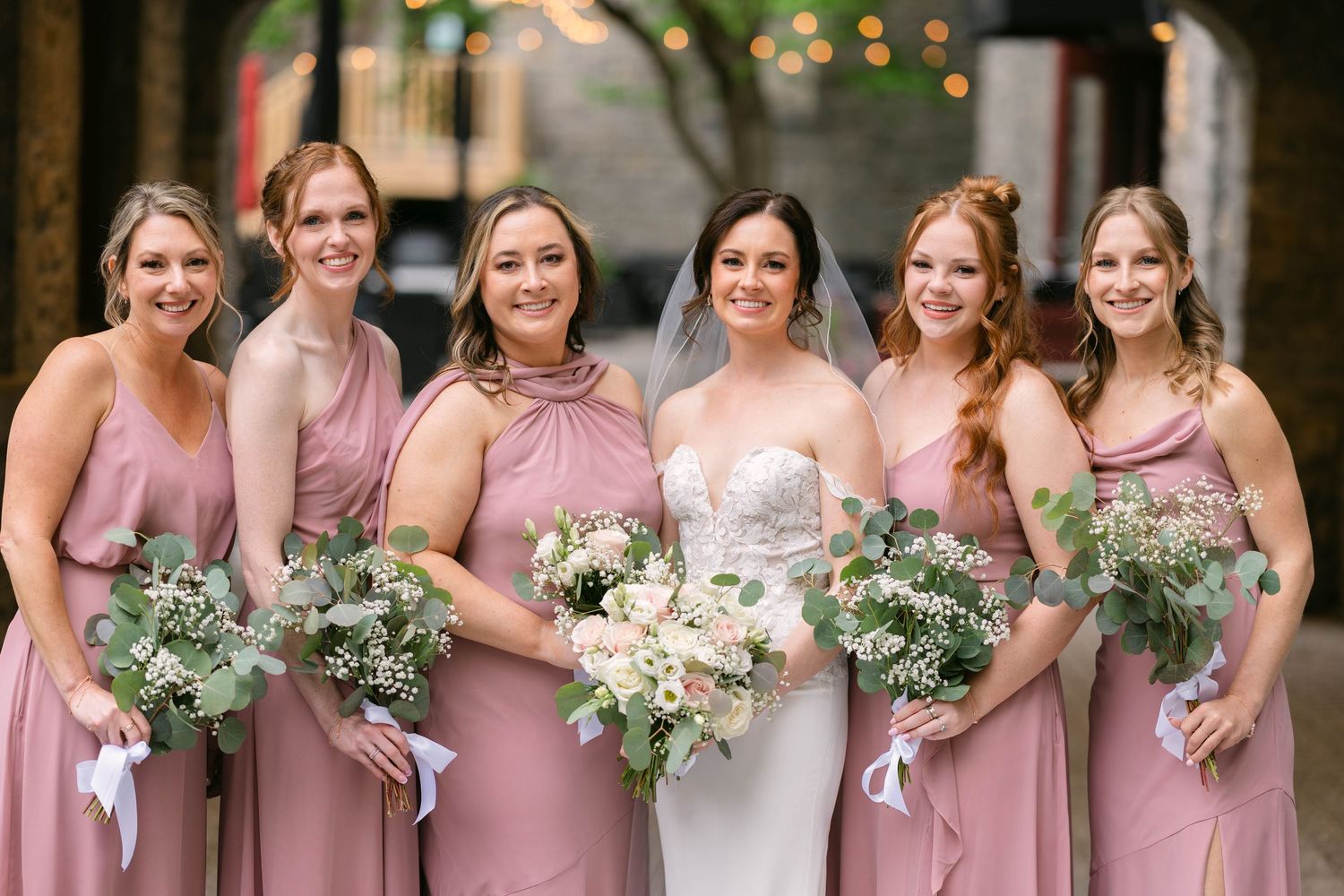 Bride in lace gown poses with five bridesmaids in dusty rose dresses holding white and greenery bouquets.