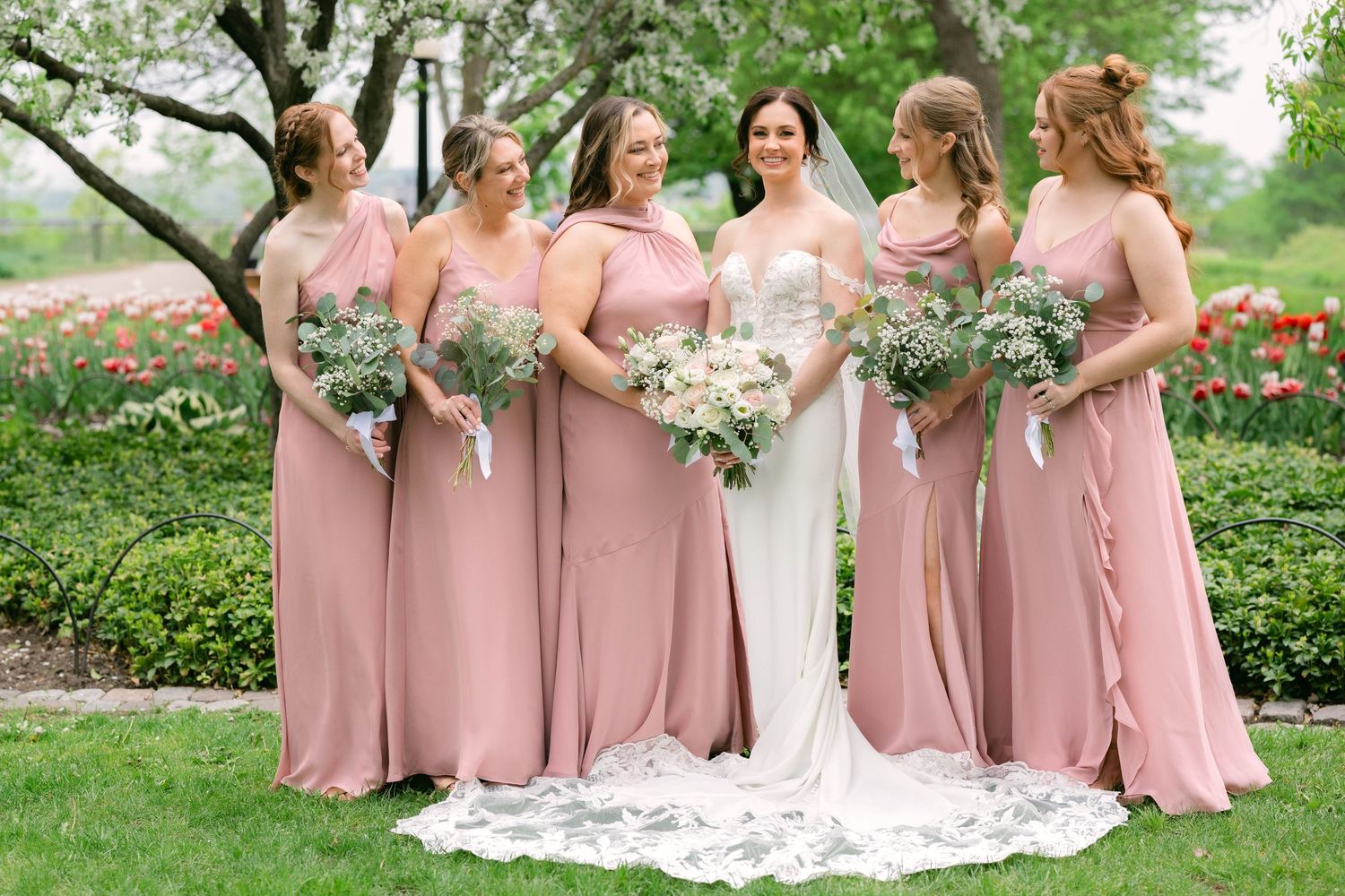 Bride in white gown with bridesmaids in dusty rose dresses holding greenery bouquets in a garden setting.