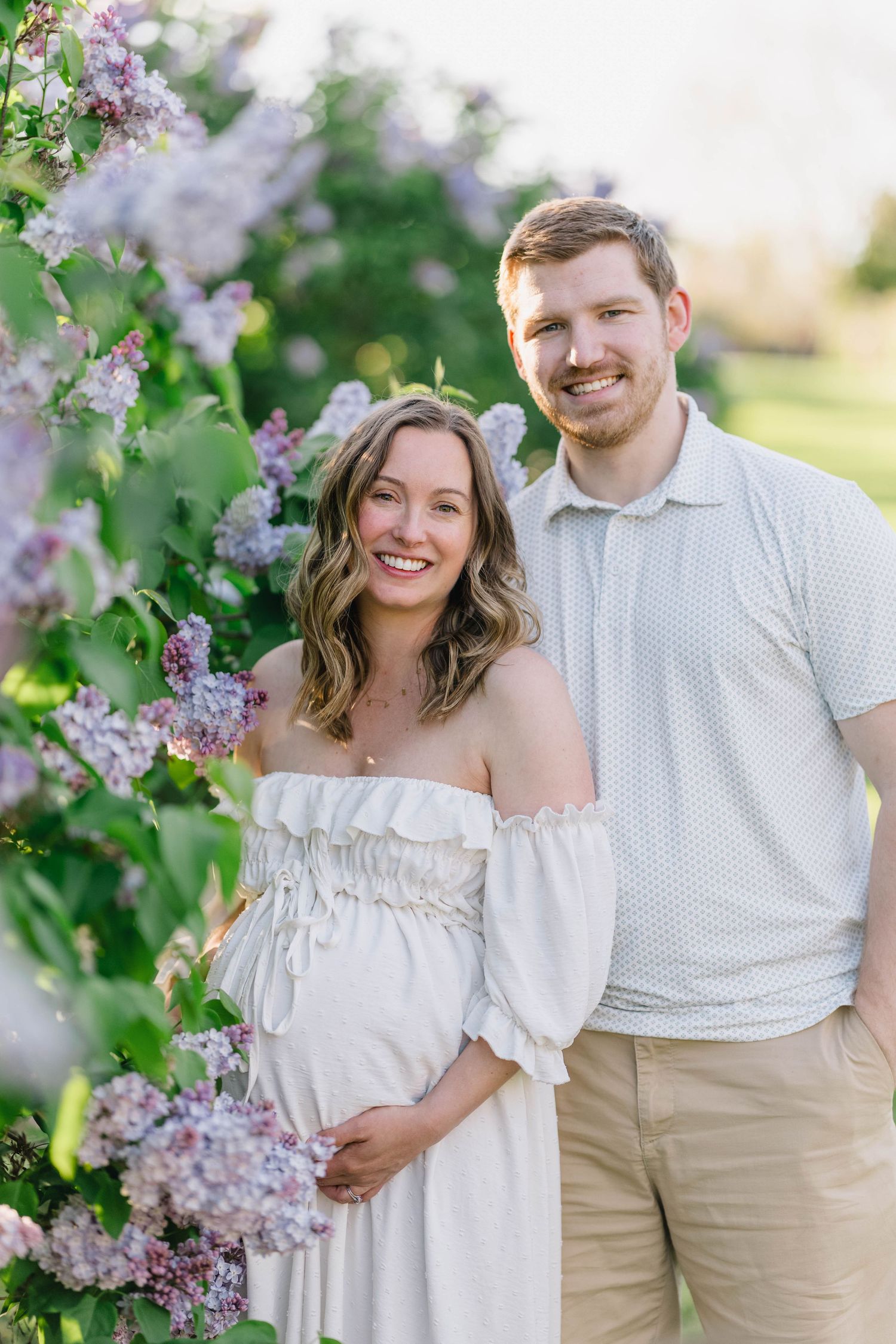 Smiling pregnant couple poses outdoors among blooming lilac bushes, woman in white off-shoulder dress, man in light polo shirt.