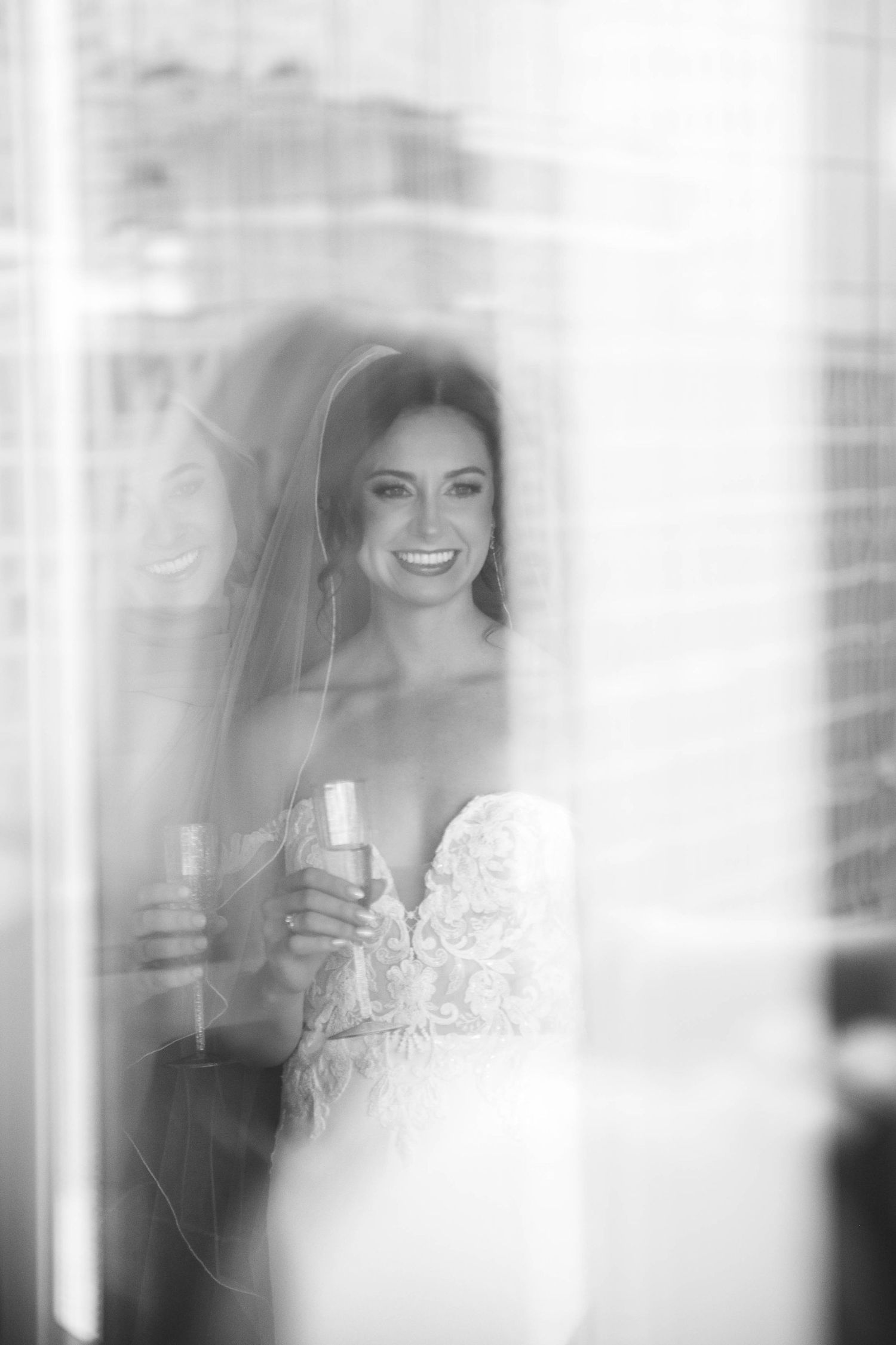 Black and white photo of a smiling bride in lace gown holding champagne glass, reflected in window light.