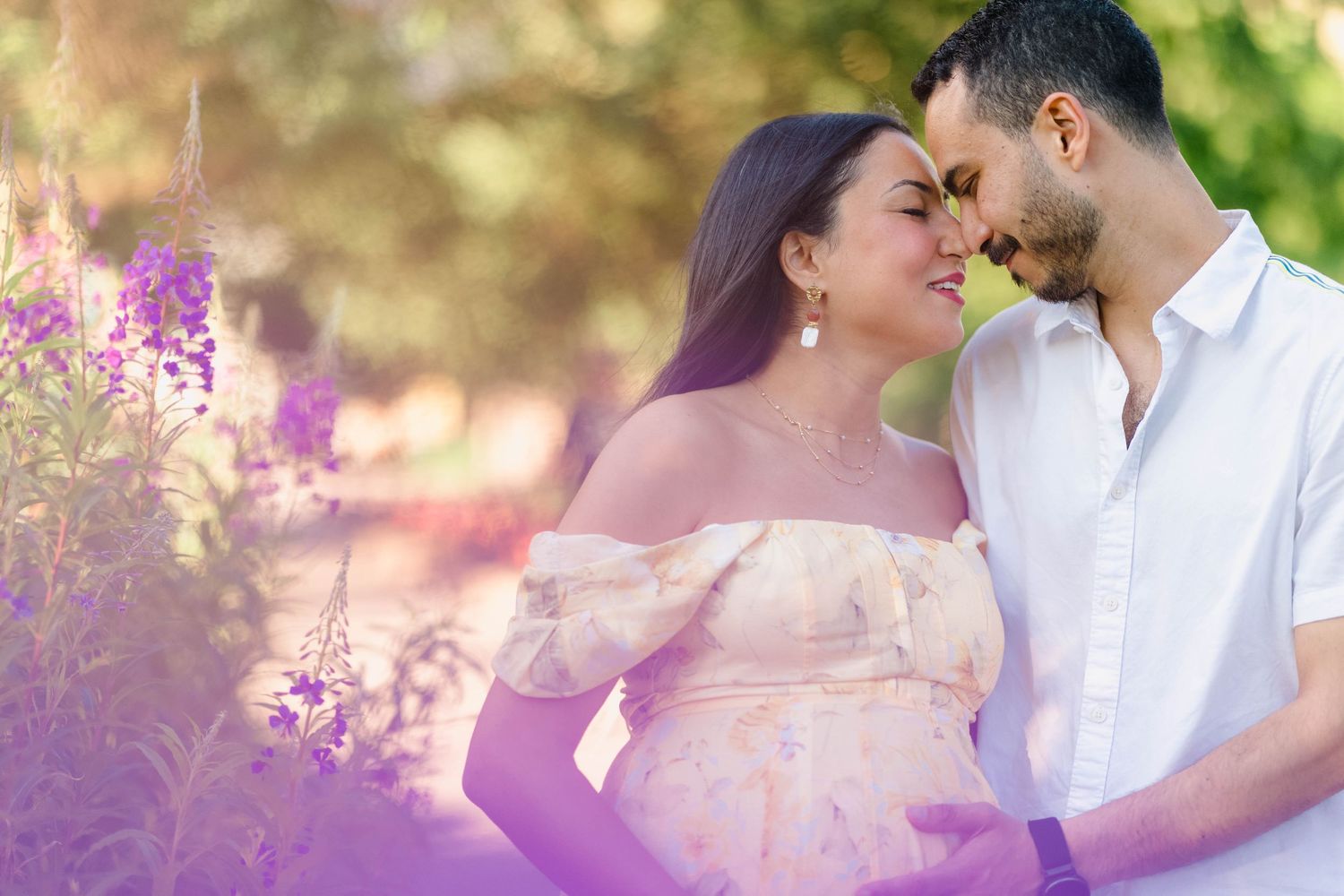 Pregnant couple sharing a tender moment outdoors surrounded by purple wildflowers in soft natural light.