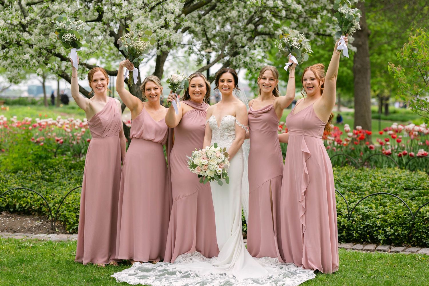 Bride and four bridesmaids in dusty rose gowns celebrate outdoors among blooming trees and colorful tulips.