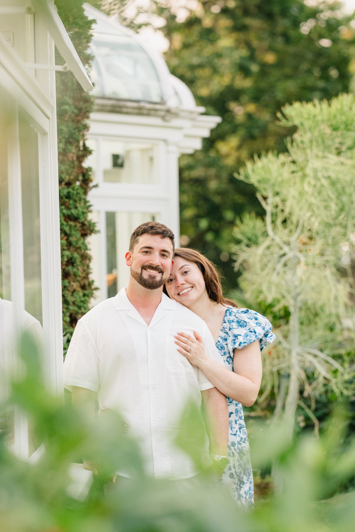 A smiling couple embraces outdoors near a white greenhouse surrounded by lush green gardens.