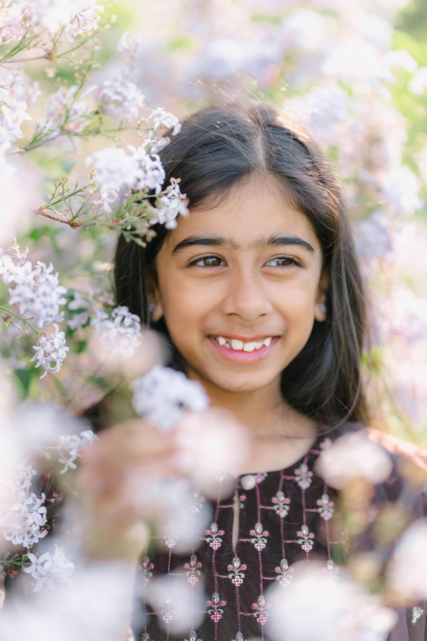 Smiling young girl with dark hair surrounded by blooming white and pink flowers in a garden.