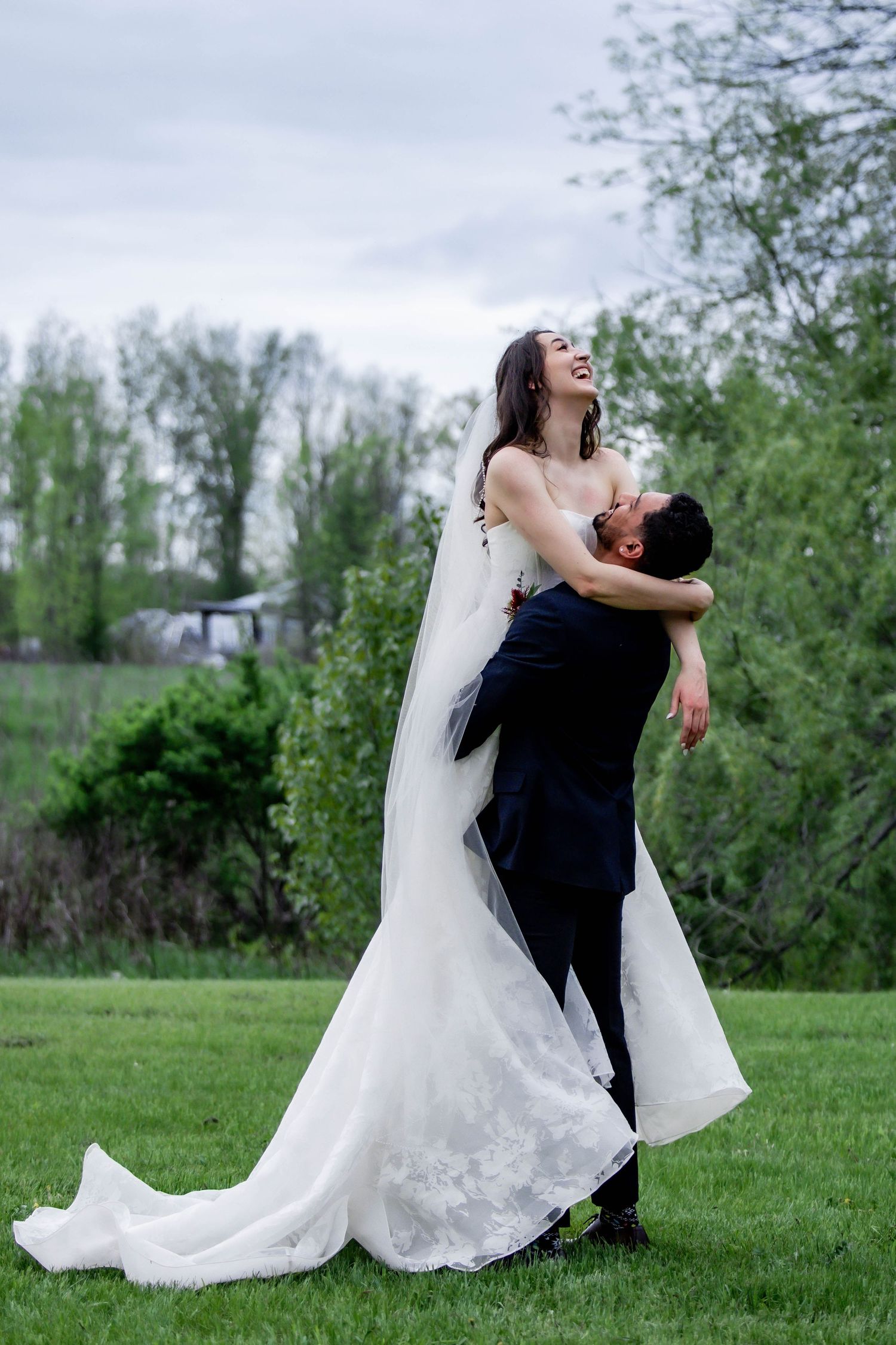 A couple in wedding attire share a joyful dance moment together on a grassy field with trees in the background.