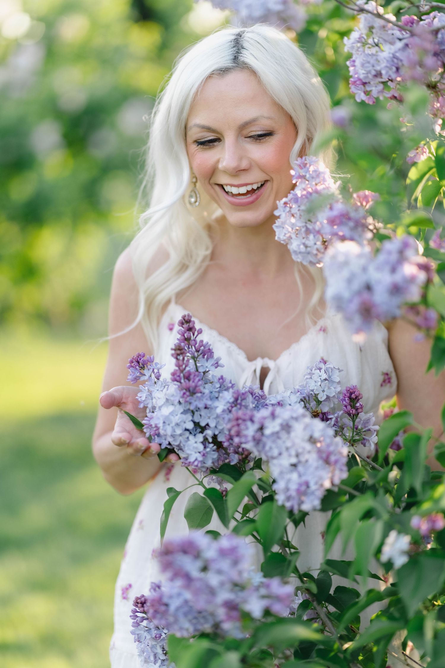 Laughing blonde woman in white dress surrounded by blooming purple lilac bushes outdoors.