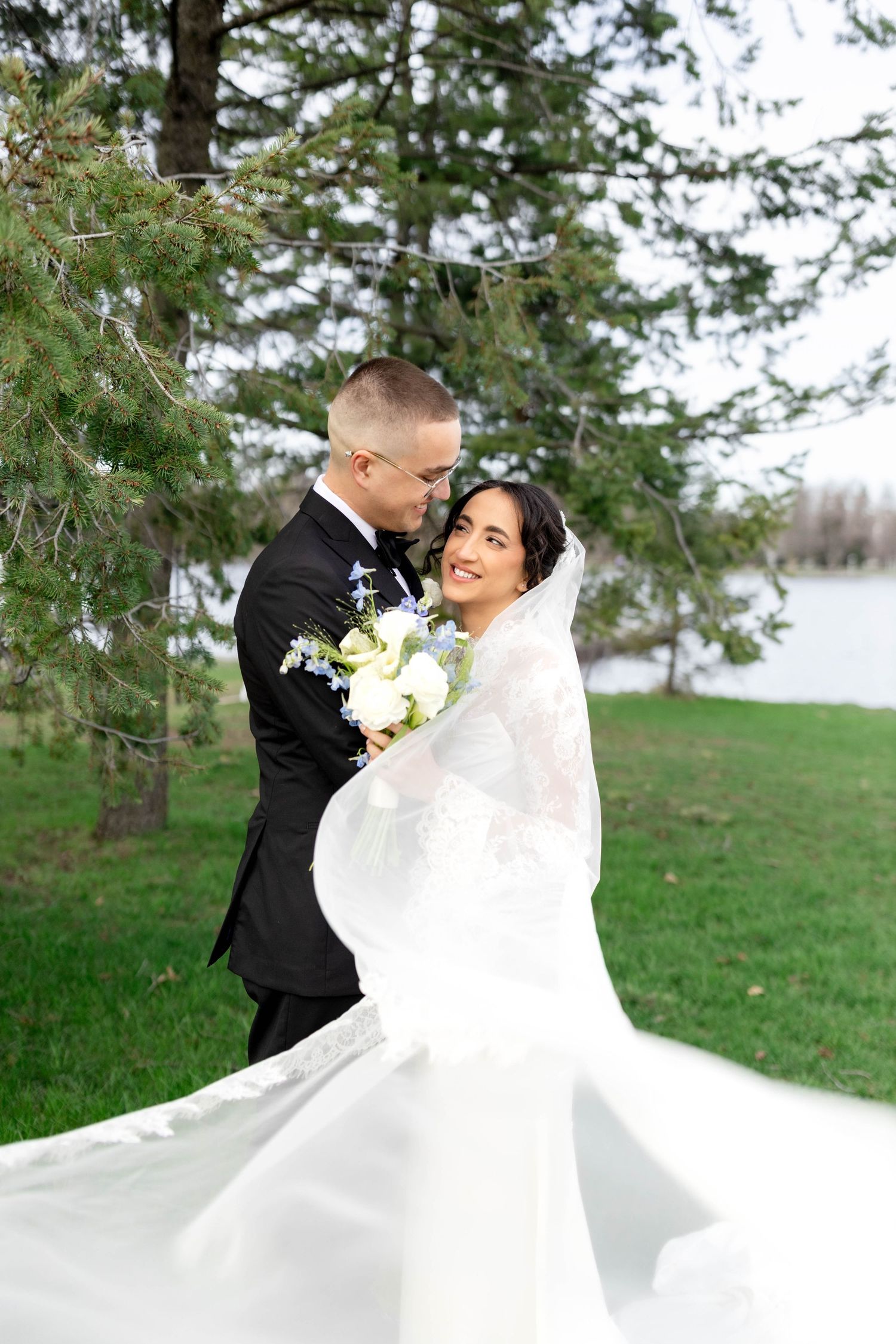 A bride and groom embrace outdoors near a pine tree and lake, her white veil flowing in the wind.