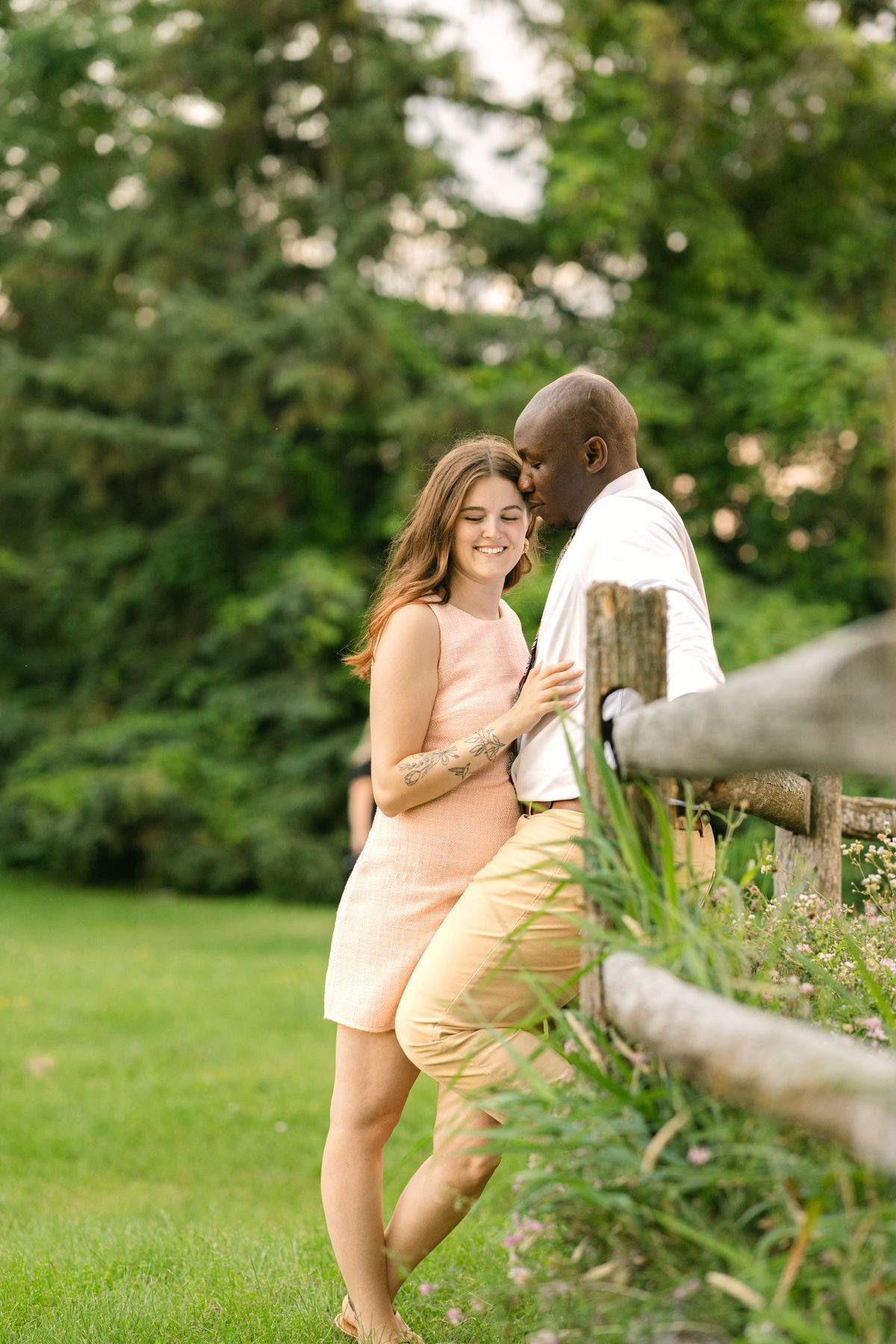 Interracial couple sharing a tender moment by a wooden fence in a lush green park, woman in pink dress smiling.