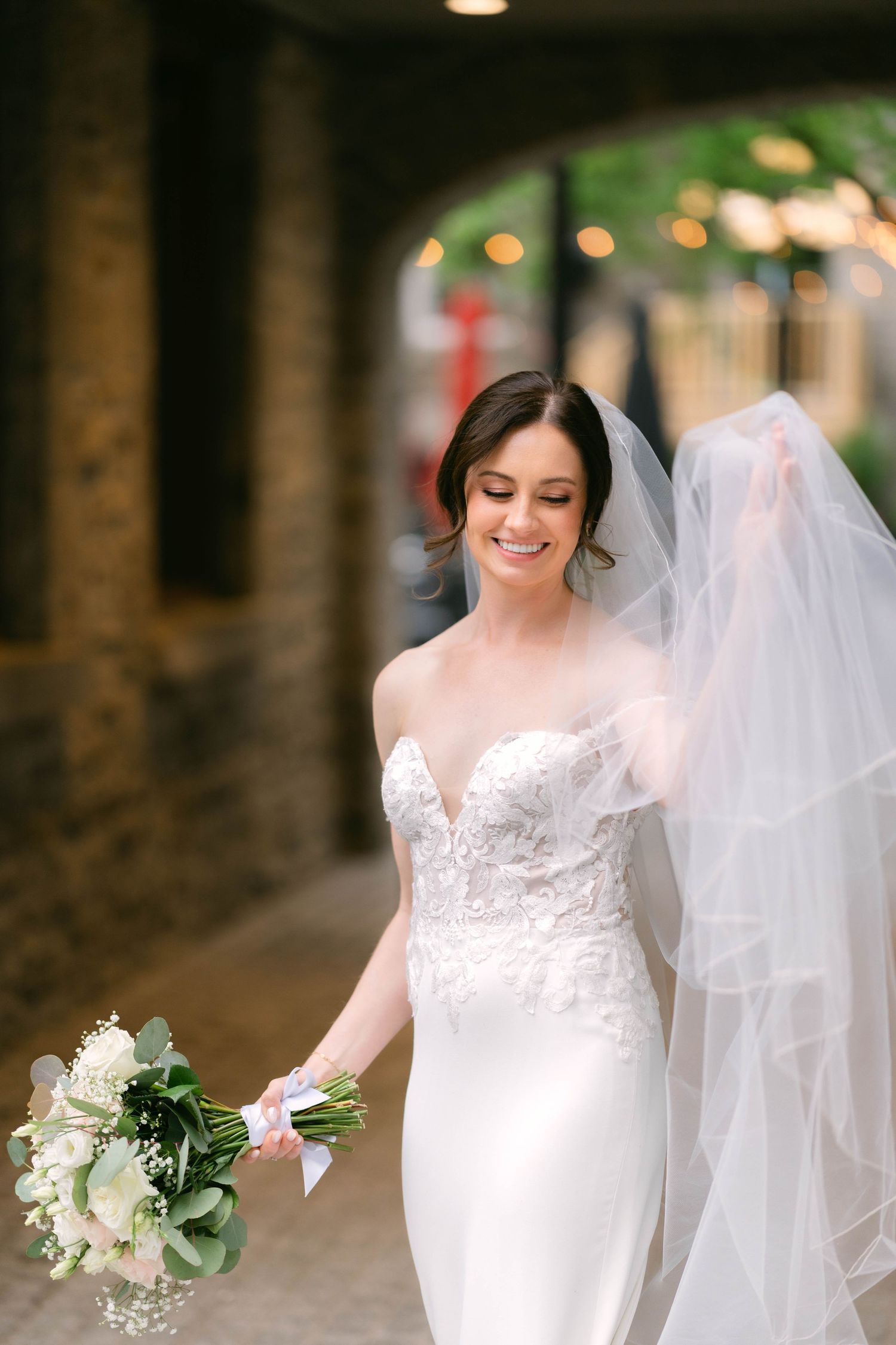 Radiant bride in strapless lace wedding dress walks with bouquet, veil flowing behind her.