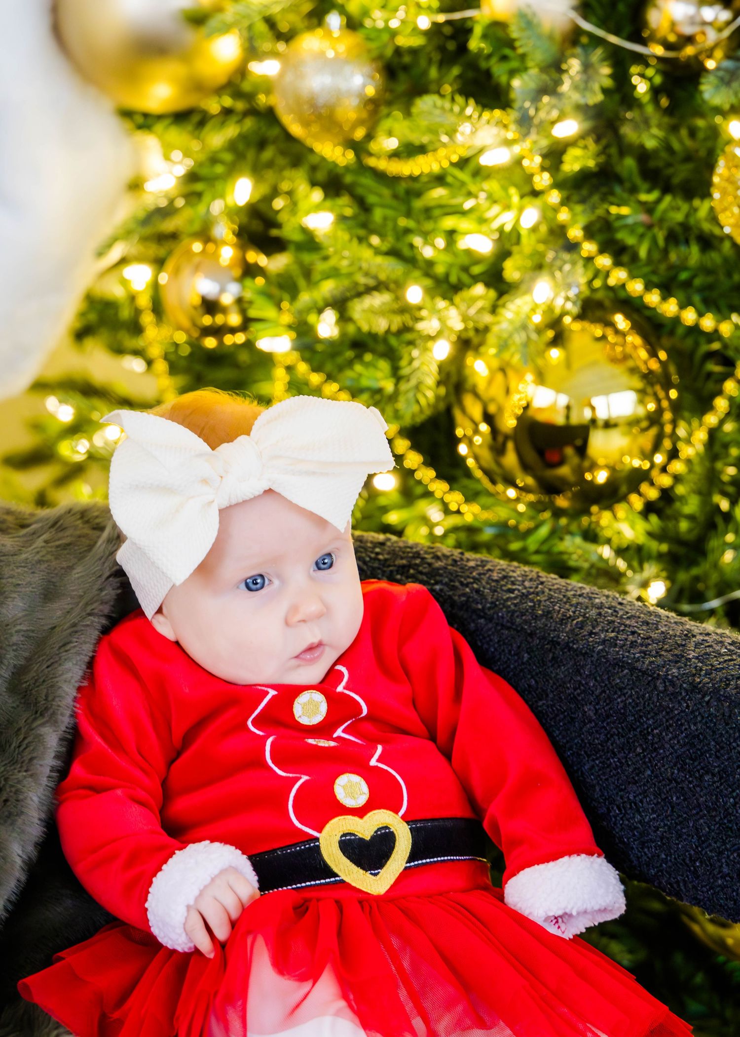 Baby wearing red Santa dress with bow near decorated Christmas tree.