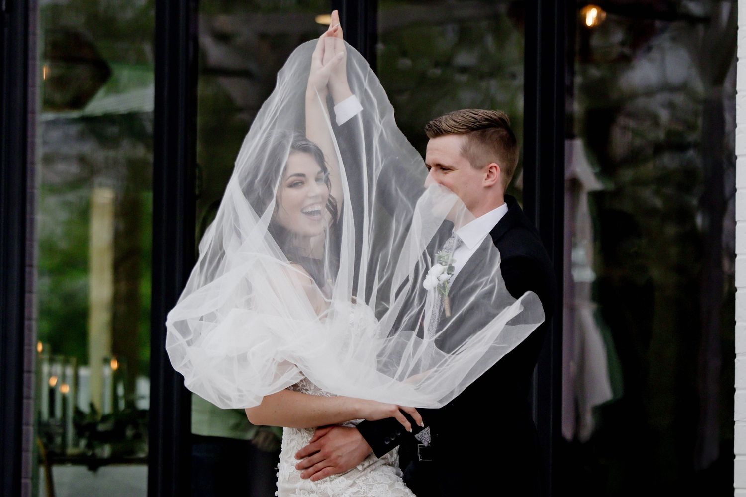 Wedding couple poses against dark windows creating a dramatic silhouette effect with the bride's veil flowing.