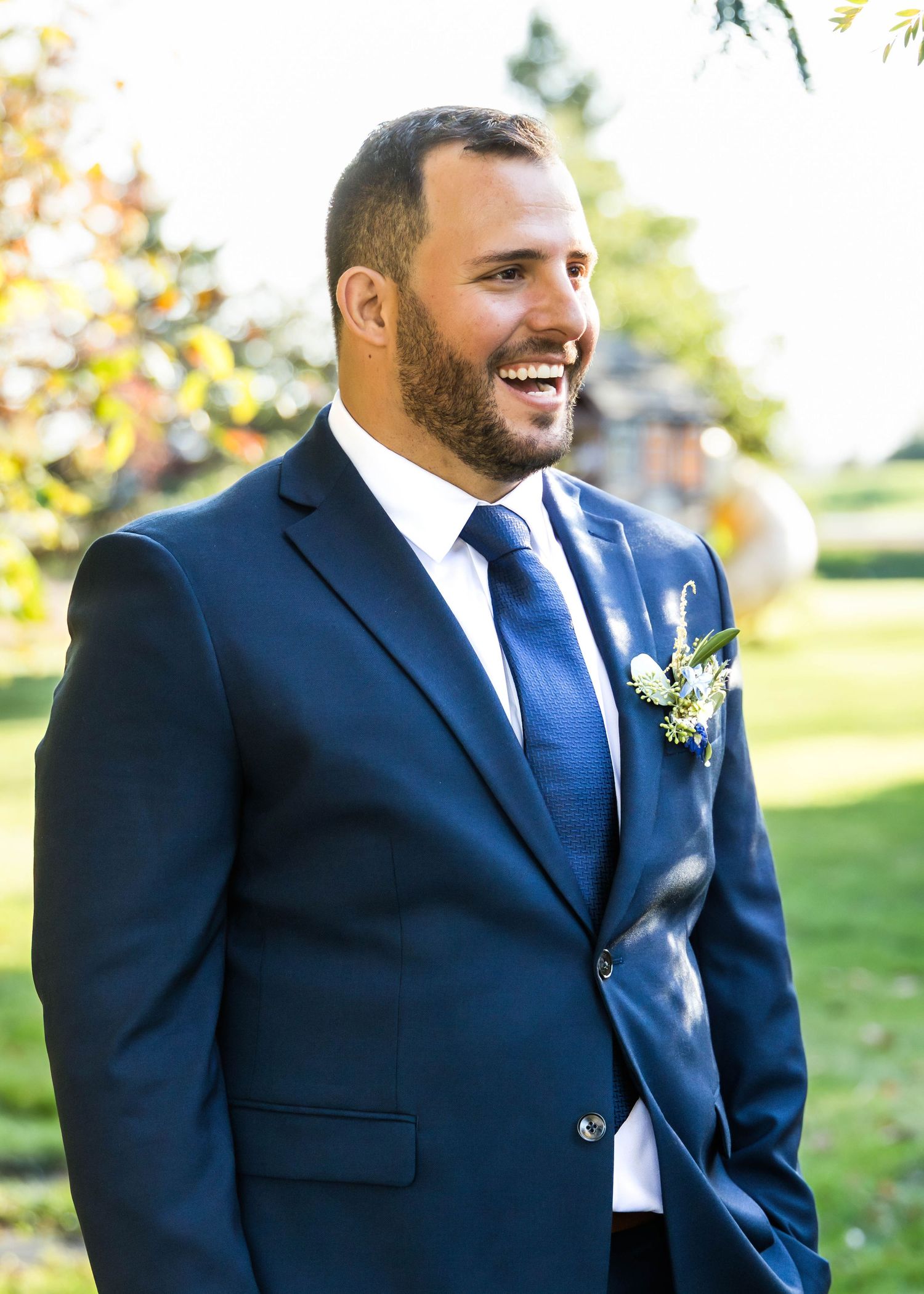 Groomsman in navy blue suit smiles while standing outdoors during autumn wedding celebration.