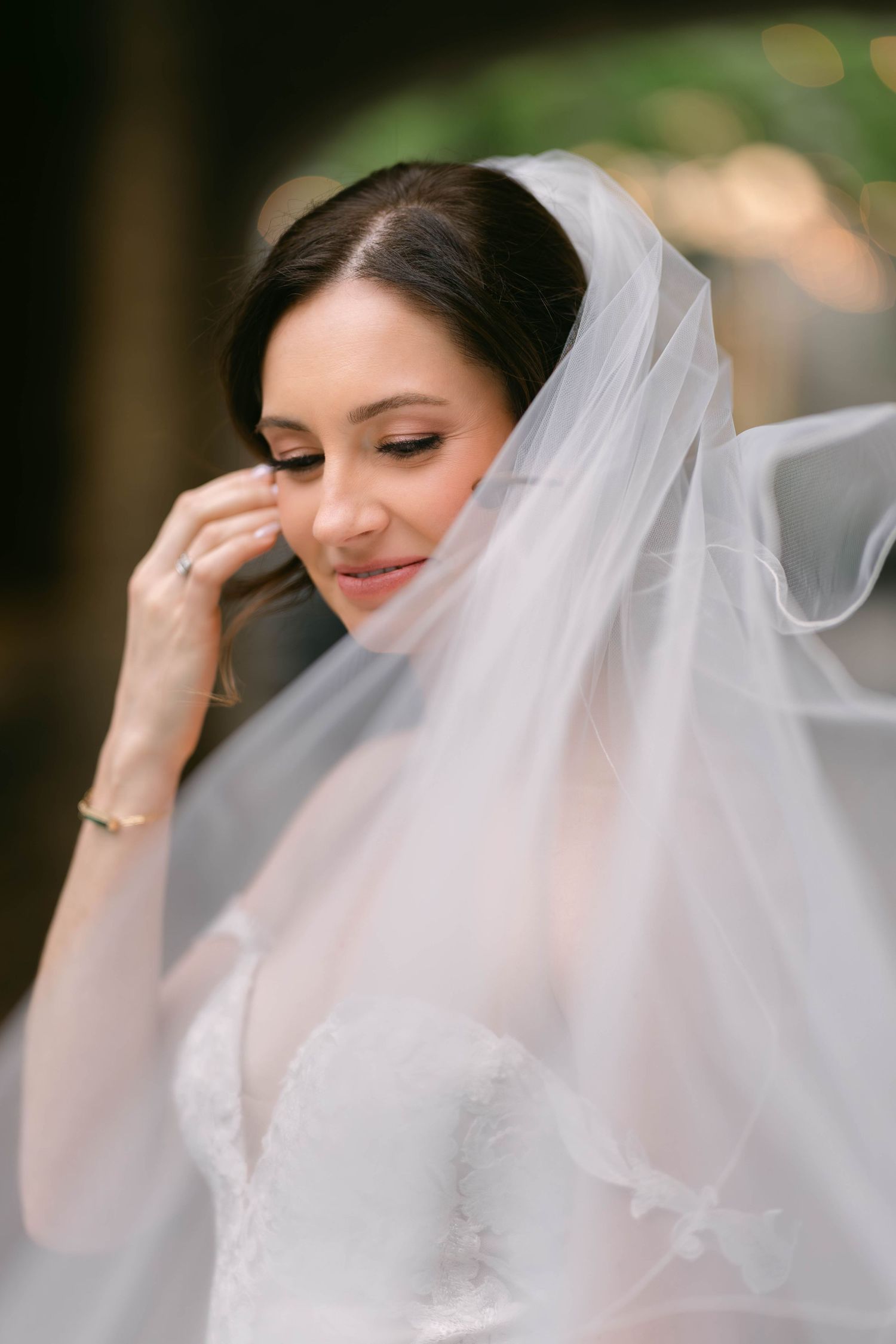 A bride in a white lace gown and flowing veil gently touches her face, eyes downcast, in a serene bridal portrait.