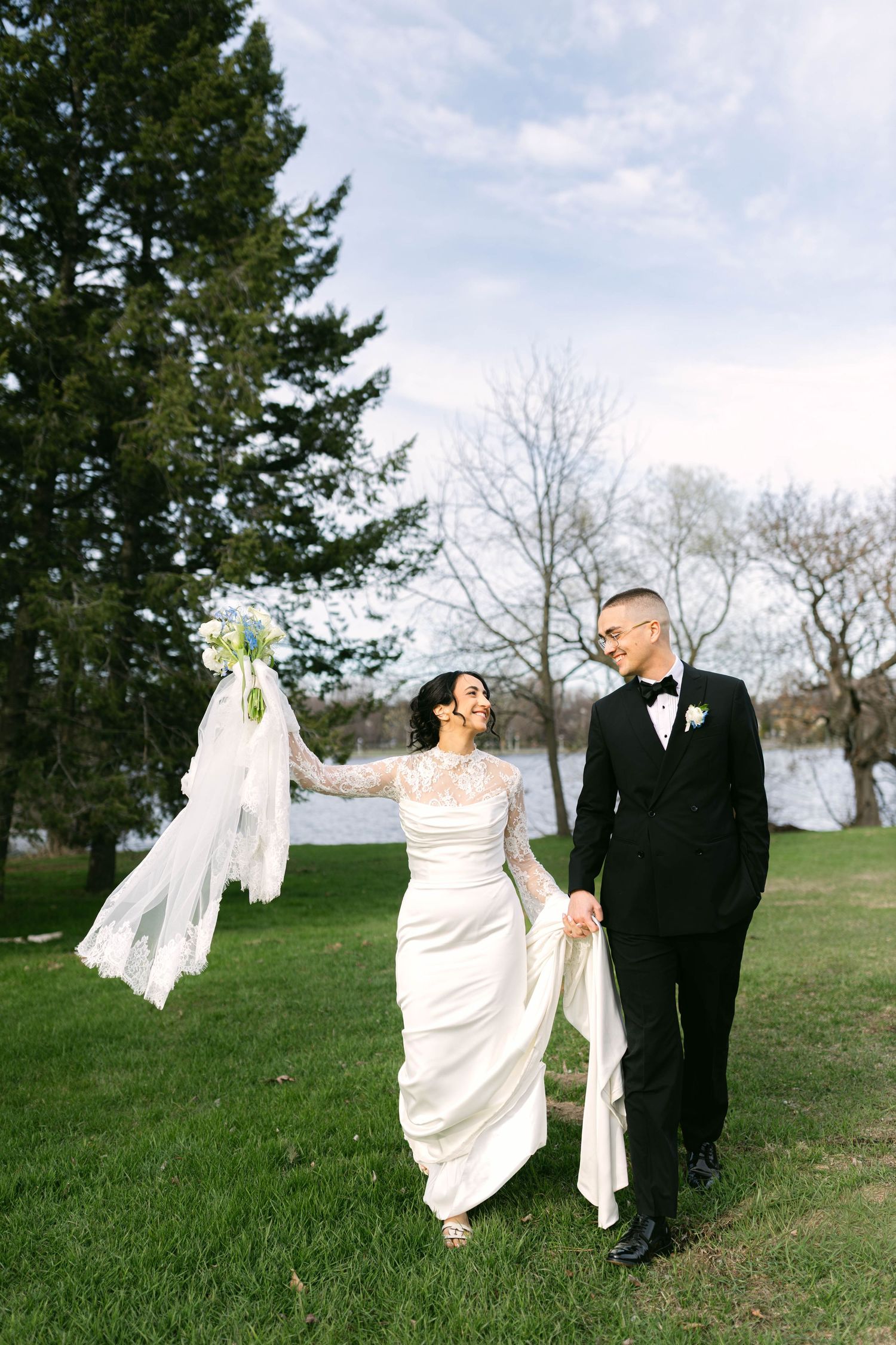 Joyful bride and groom walk hand in hand on green lawn, bride raises bouquet and veil in the air.