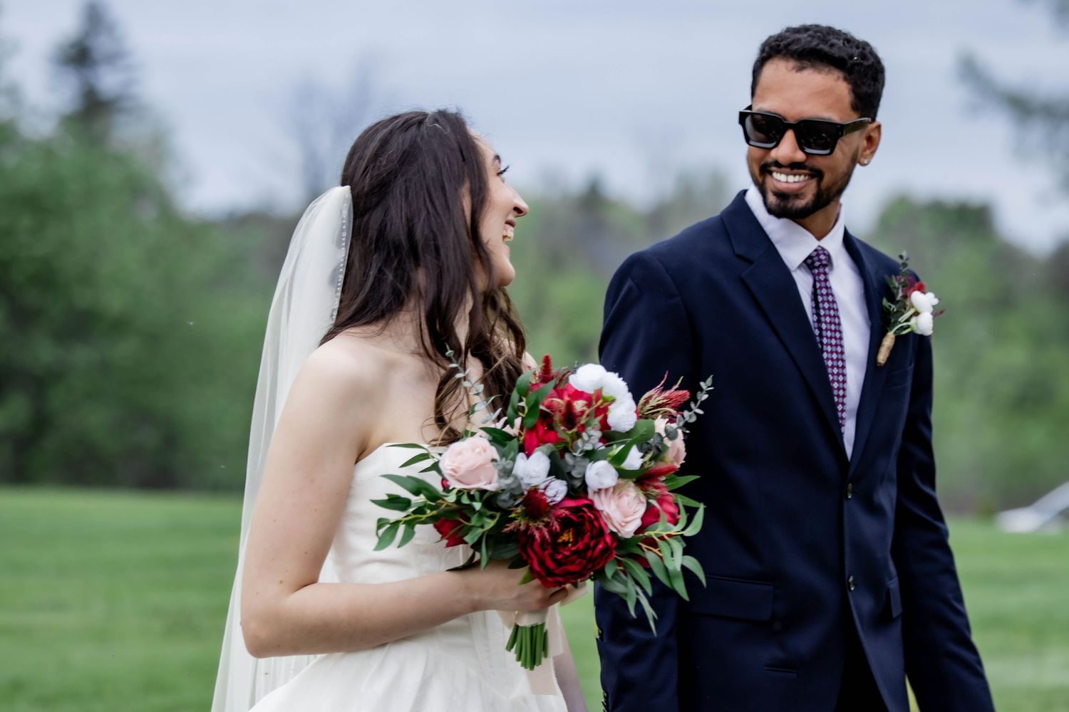Outdoor wedding scene with couple holding red rose bouquet against lush green landscape.