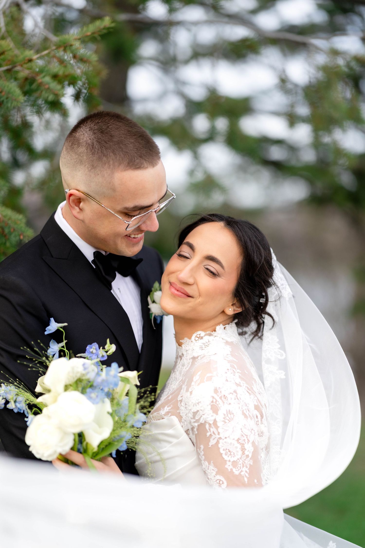 Bride and groom embrace outdoors, she holds blue and white bouquet, wearing lace gown with veil, he wears black tuxedo.