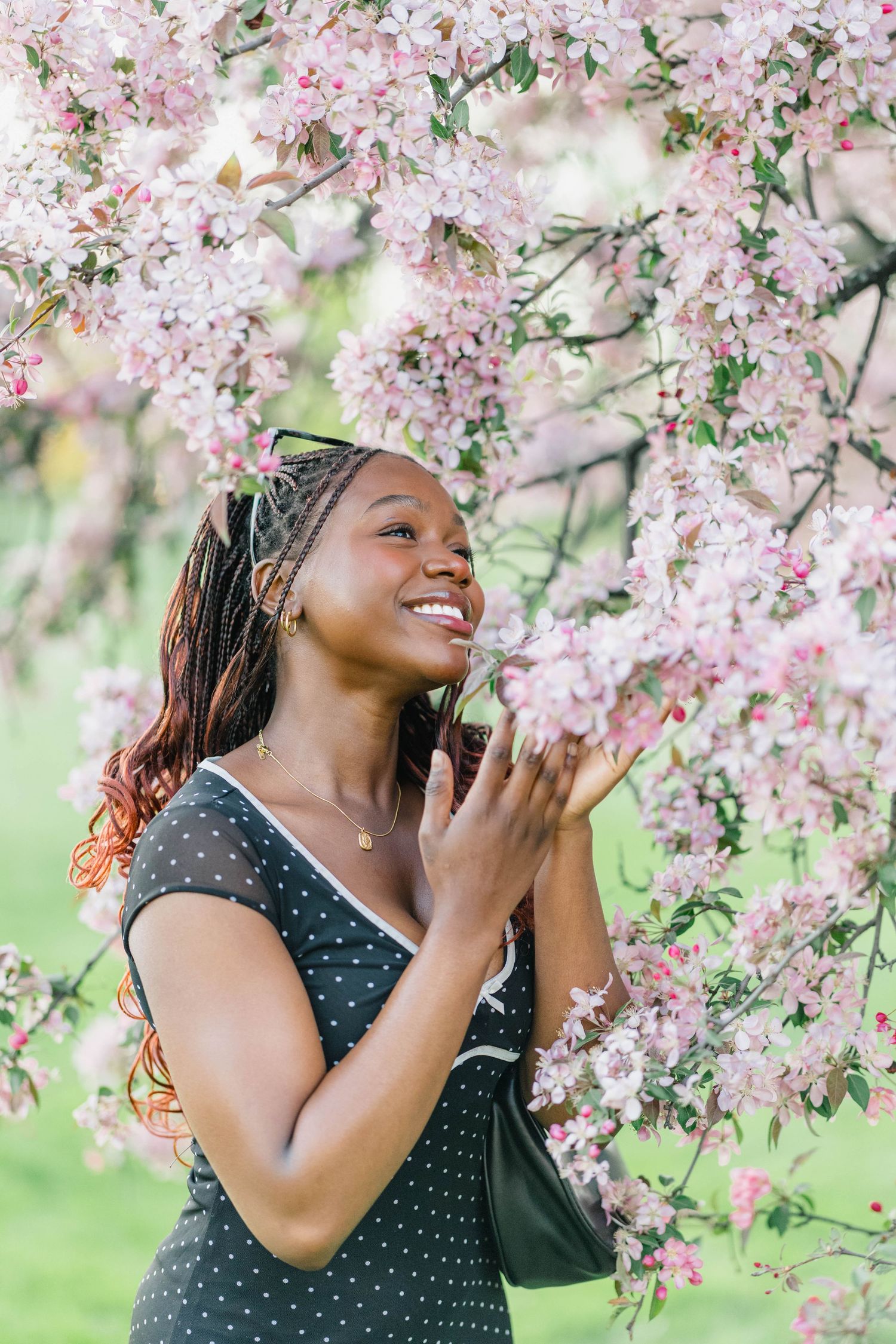 A smiling young Black woman in a polka dot dress enjoys pink cherry blossoms in a lush green garden.